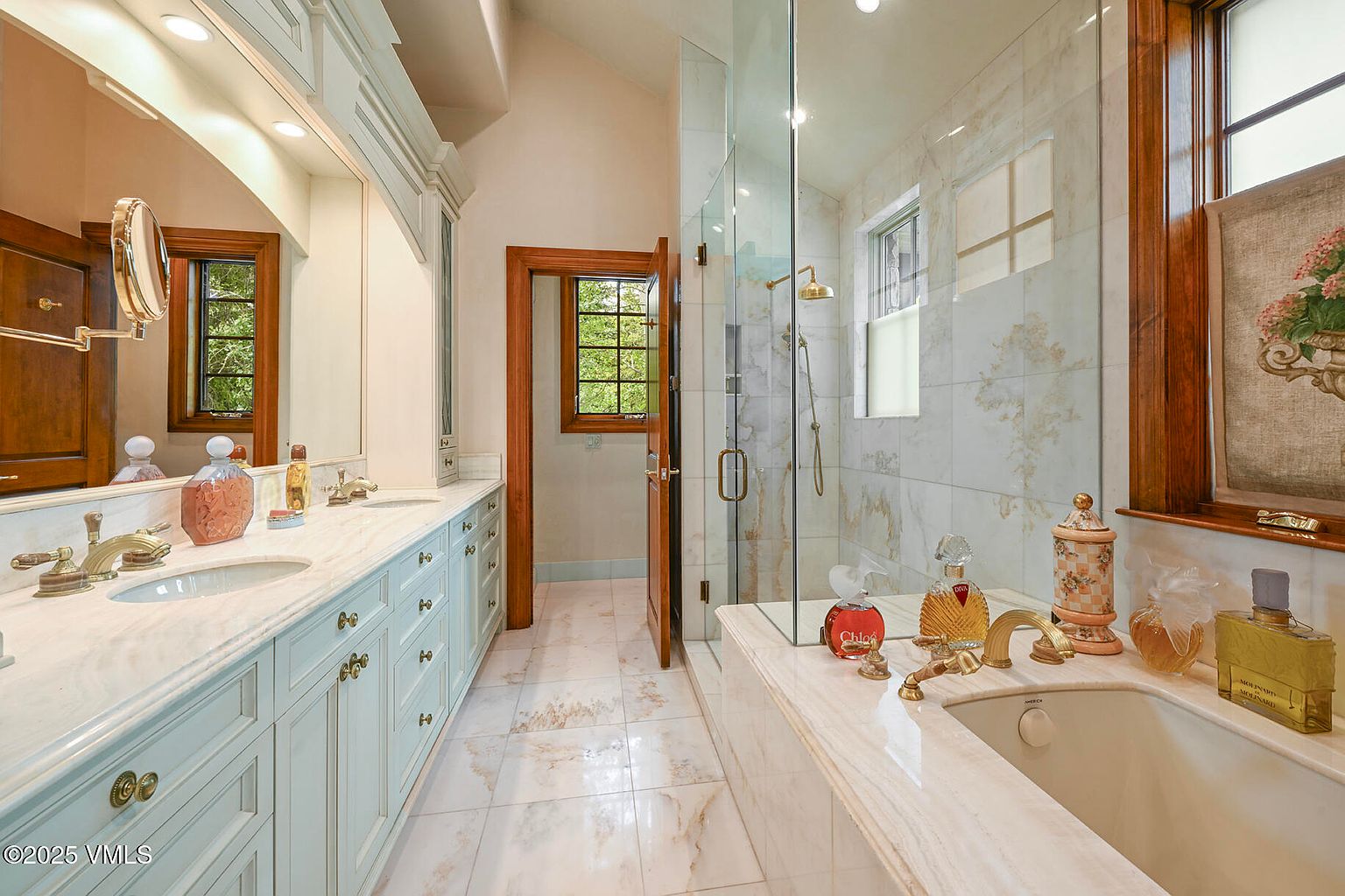 This is a luxurious primary bathroom featuring a double vanity with light blue cabinetry and marble countertops. A glass-enclosed shower is adjacent to a soaking tub, both adorned with gold fixtures. The room is well-lit and exudes a spa-like ambiance with decorative perfume bottles and a window providing natural light.