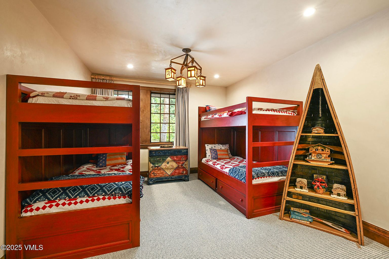 This is a cozy guest bedroom featuring two sets of red-stained wooden bunk beds, creating a rustic and inviting atmosphere. A decorative chest sits beneath a window with natural light, and a unique boat-shaped shelving unit adds character to the room. The neutral-toned carpet and walls provide a calming backdrop to the colorful bedding and decor.