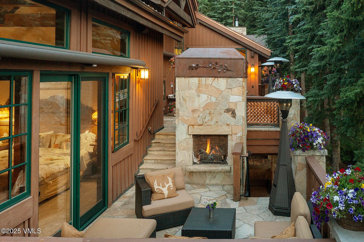 This image showcases a charming outdoor patio area, complete with a stone fireplace and comfortable seating. The patio features a stone floor and is surrounded by lush greenery, creating a cozy and inviting atmosphere. The presence of outdoor heaters suggests the space is designed for year-round enjoyment.