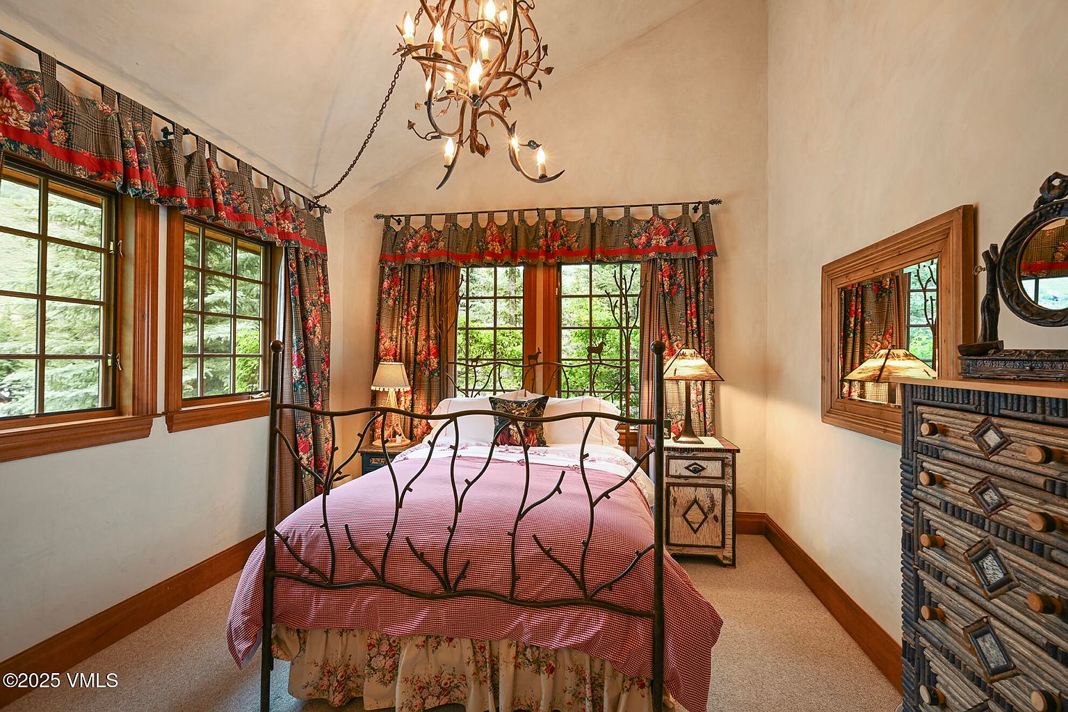 This is a cozy bedroom featuring a wrought iron bed with red and white striped bedding and a floral bed skirt. The room has a rustic charm with wooden window frames, floral curtains, and a unique chandelier. A wooden dresser with a mirror adds to the room's character.