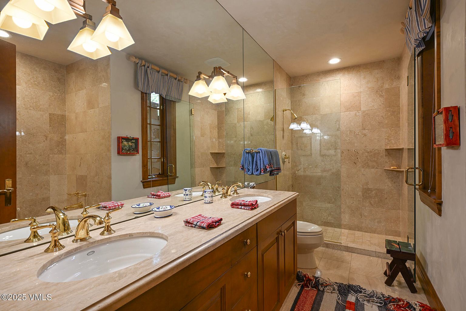 This is a well-lit primary bathroom featuring a double vanity with a light-colored countertop and wooden cabinetry. A large mirror spans the length of the vanity, reflecting the lighting fixtures above. The bathroom includes a glass-enclosed shower and is tiled in a neutral tone, creating a clean and luxurious feel.