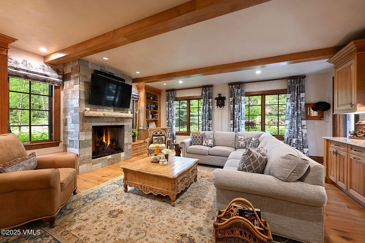 This is a warm and inviting living room featuring a stone fireplace with a mounted television above. The room is furnished with a comfortable sectional sofa, an armchair, and a decorative wooden coffee table, all set upon an ornate area rug. Natural light streams in through the windows, complemented by wooden ceiling beams, creating a cozy and elegant atmosphere.