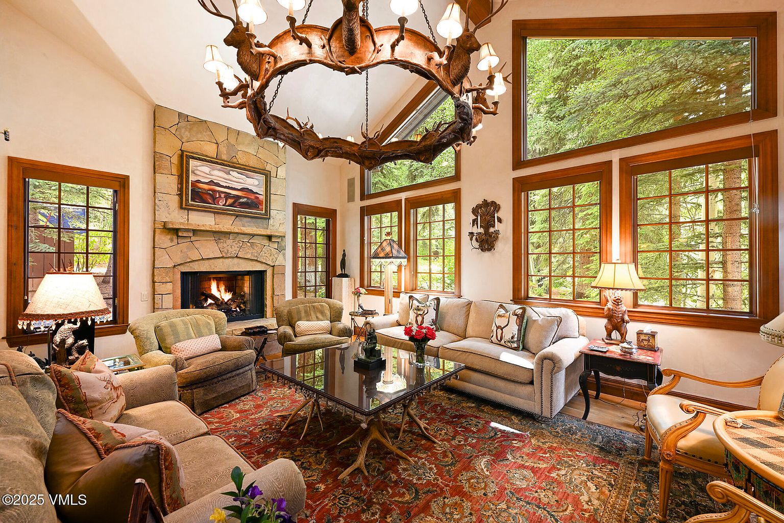 This is a warm and inviting living room featuring a stone fireplace, a unique antler chandelier, and large windows that offer views of the surrounding trees. The room is furnished with comfortable seating, including sofas and armchairs, arranged around a glass-topped coffee table on an ornate rug. The overall style is rustic and luxurious, creating a cozy and elegant atmosphere.