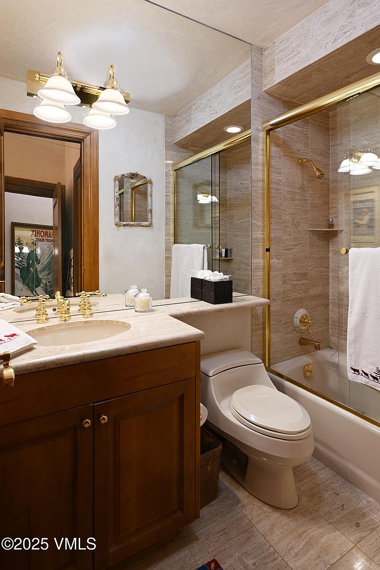 This is a well-lit bathroom featuring a wooden vanity with a light-colored countertop and gold fixtures. A toilet is positioned next to the vanity, and a shower-tub combination with glass doors and gold trim is visible. The walls and floor are tiled in a neutral tone, creating a clean and classic look.