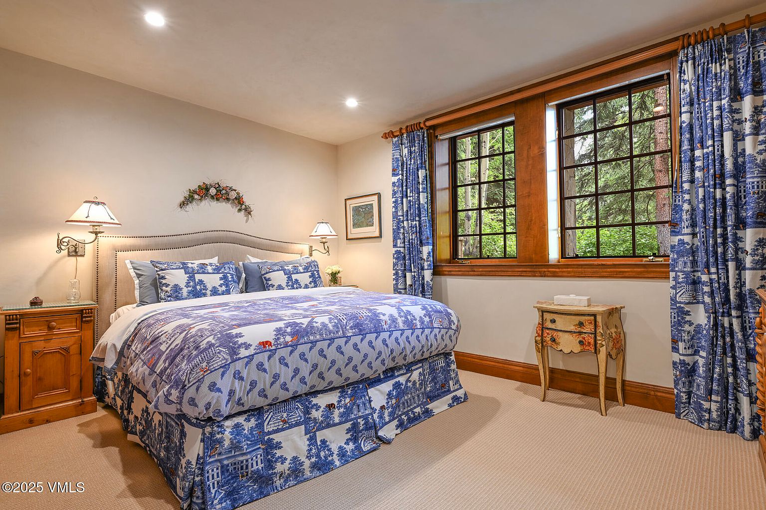 This is a bedroom featuring a bed with blue and white patterned bedding, flanked by wooden nightstands and lamps. The room has two windows with matching blue and white curtains, providing natural light. The overall style is traditional and cozy, creating a comfortable and inviting atmosphere.