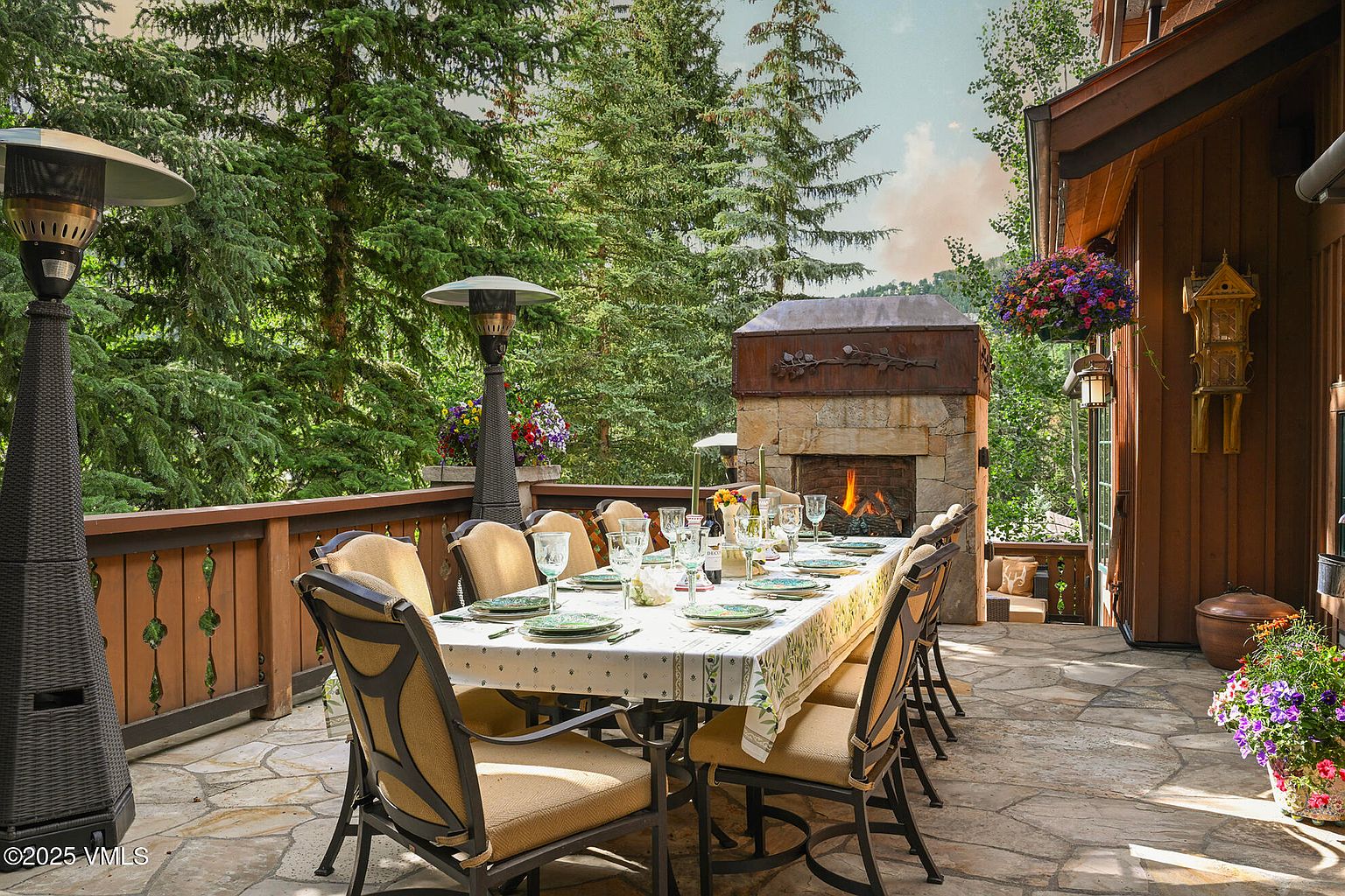 This image showcases a beautifully arranged outdoor dining area on a stone patio. A long table is set for a meal, surrounded by comfortable chairs, with a stone fireplace providing a warm ambiance. The space is framed by lush greenery and trees, creating a serene and inviting atmosphere, perfect for entertaining or relaxing outdoors.