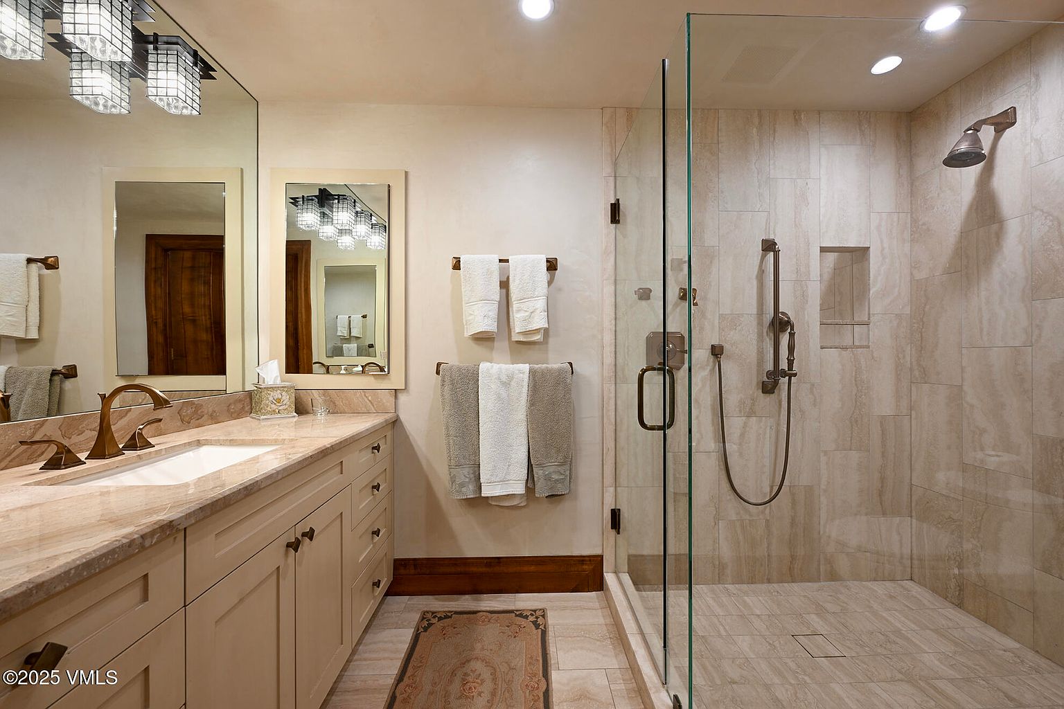 This is a well-lit primary bathroom featuring a double vanity with a marble countertop and light-colored cabinetry. A large mirror and decorative lighting fixtures are above the vanity. The shower is enclosed with glass and has travertine tile walls and floor, with bronze fixtures. The overall impression is clean, elegant, and luxurious.