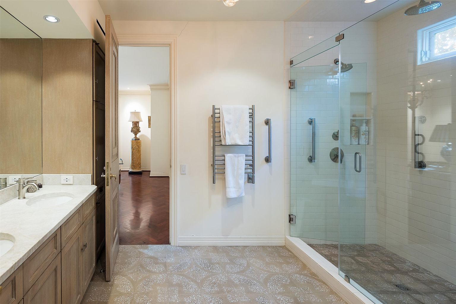 This bright bathroom features a double vanity with light wood cabinetry and a stone countertop, complemented by a large mirror. To the right, a glass-enclosed walk-in shower with white subway tile walls and a patterned tile floor creates a clean, modern aesthetic. A wall-mounted heated towel rack with white towels adds a functional touch, while an open doorway provides a view into an adjacent room with dark wood flooring.