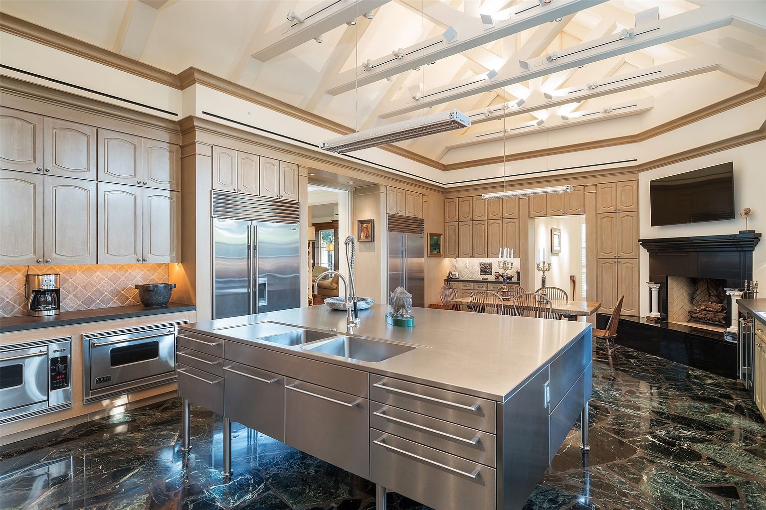 This expansive, high-end kitchen features a massive stainless steel central island with an integrated sink, set against a backdrop of light wood cabinetry and dark stone flooring. The room is defined by a dramatic vaulted ceiling with exposed white beams and professional-grade stainless steel appliances, including dual refrigerators and wall ovens. A black fireplace surround adds a striking focal point to the space, which flows seamlessly into a formal dining area, creating an atmosphere of sophisticated luxury.