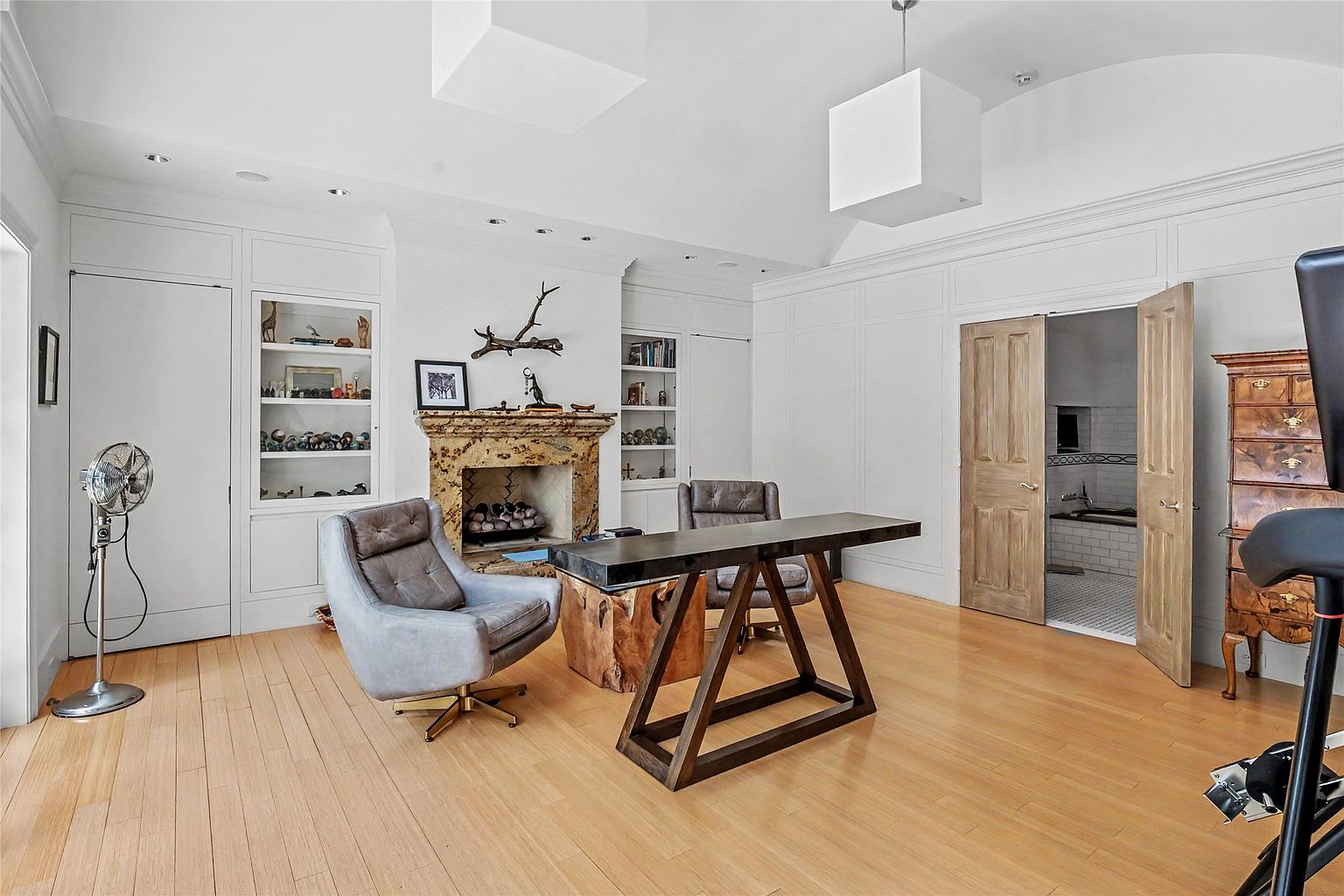 This sophisticated home office features light wood flooring and a prominent stone fireplace with a decorative mantel. The room is furnished with a unique desk supported by a natural wood base and two grey leather armchairs, while built-in shelving units flank the fireplace. Double wooden doors open into an adjacent room, and the space is illuminated by modern, box-shaped pendant lights.
