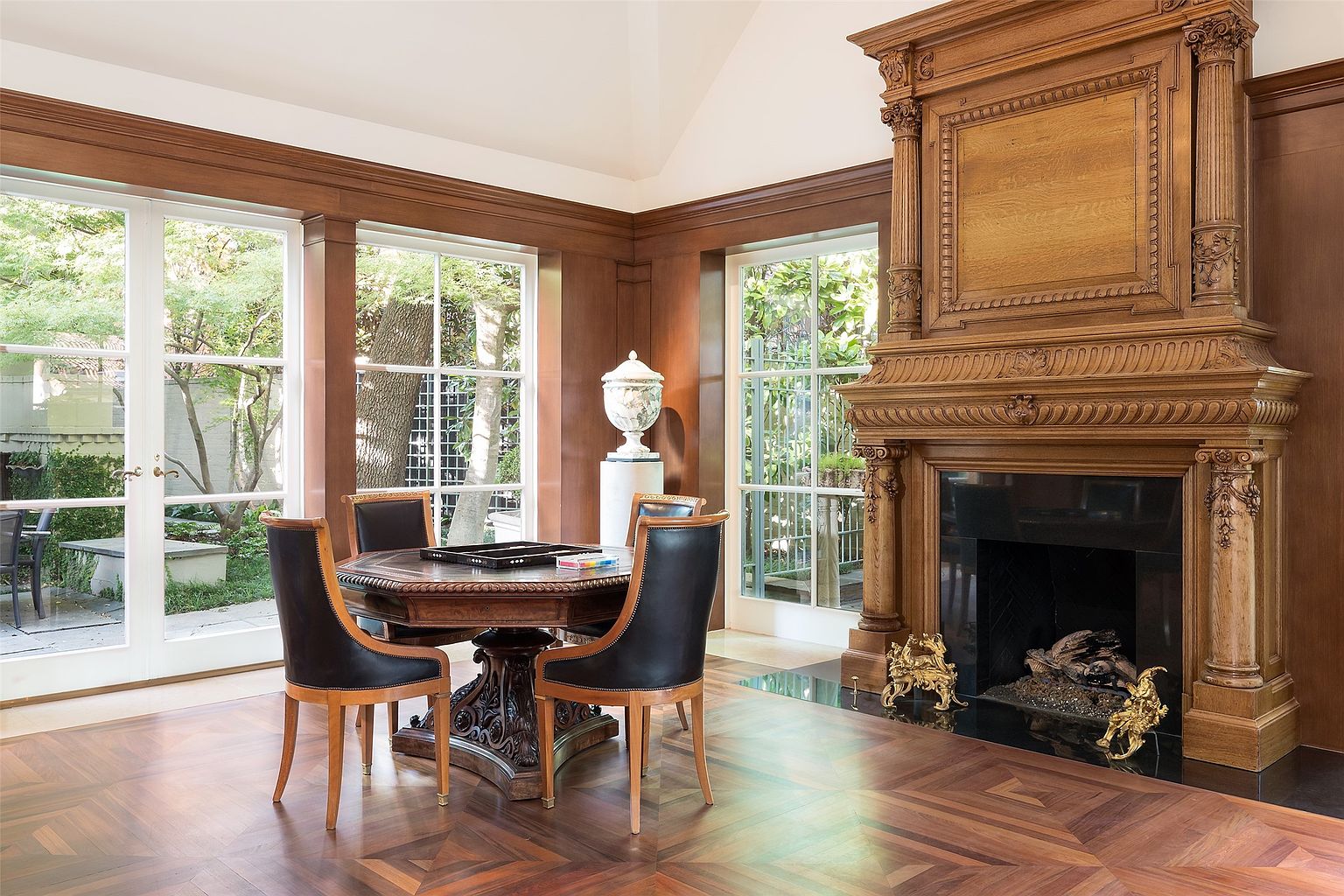 This elegant room features a prominent, intricately carved wood fireplace surround that serves as a focal point against the wood-paneled walls. A circular game table with four leather-upholstered chairs sits in the center of the room, set upon a rich, patterned wood floor. Large windows and glass doors provide abundant natural light and views of the lush greenery outside, creating a sophisticated and inviting atmosphere.