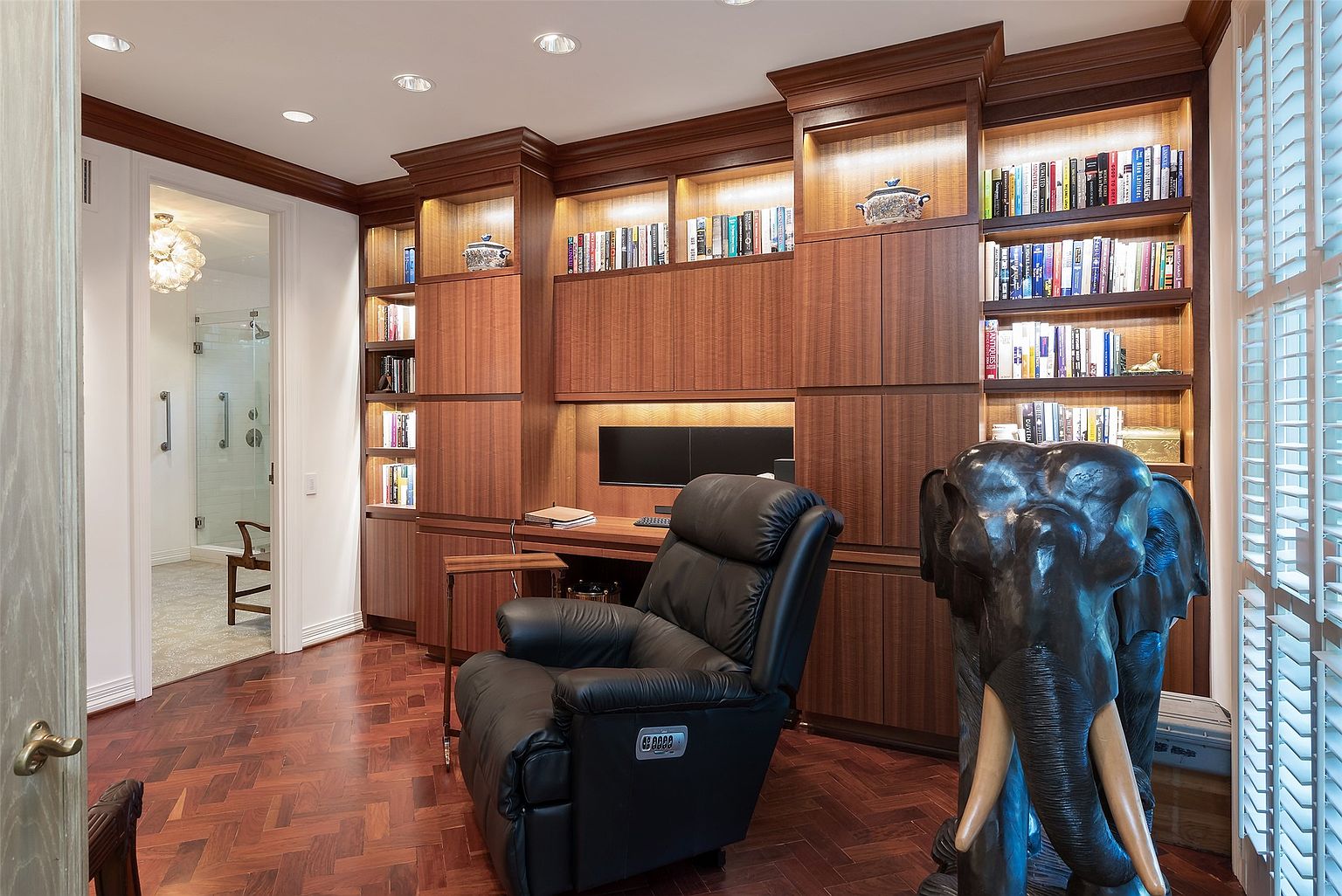 This sophisticated home office features rich, dark wood built-in cabinetry with integrated lighting that highlights the shelving and desk area. A plush black leather recliner sits in the center of the room, positioned on a classic herringbone-patterned wood floor. To the right, a large, detailed elephant sculpture adds a unique decorative touch, while a doorway on the left provides a glimpse into a bright, tiled bathroom.