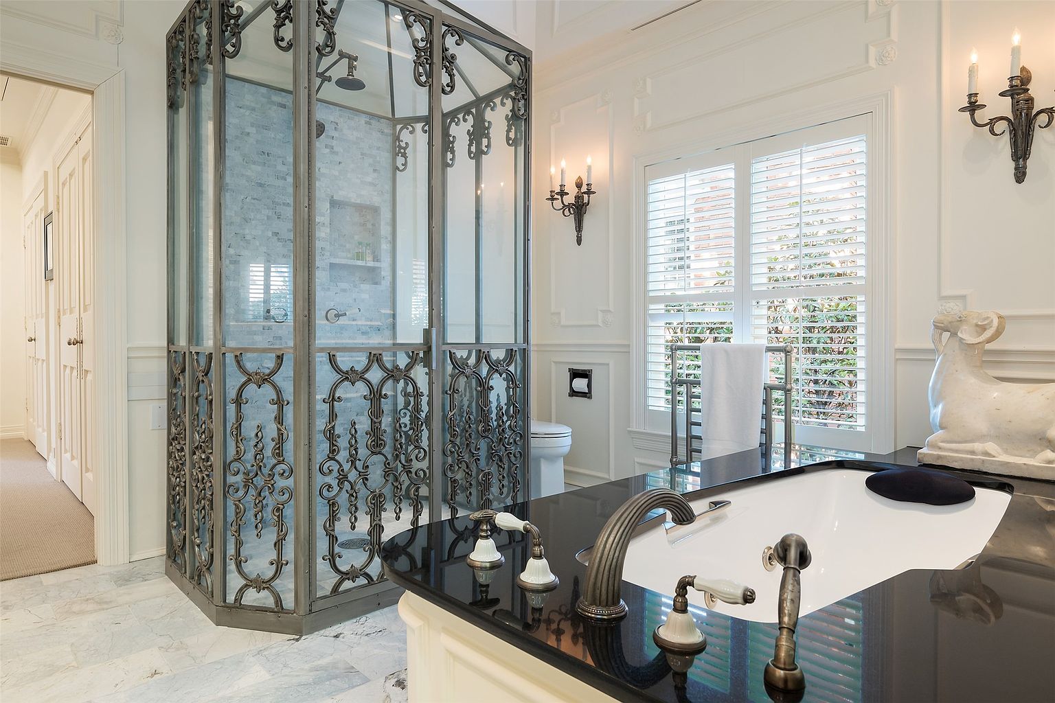 This elegant primary bathroom features a striking custom shower enclosure with intricate metal scrollwork and glass panels. A dark stone-topped vanity with a classic faucet sits in the foreground, complemented by a decorative ram sculpture and wall-mounted candle-style sconces. The space is finished with light-toned wall paneling and plantation shutters, creating a sophisticated and luxurious atmosphere.