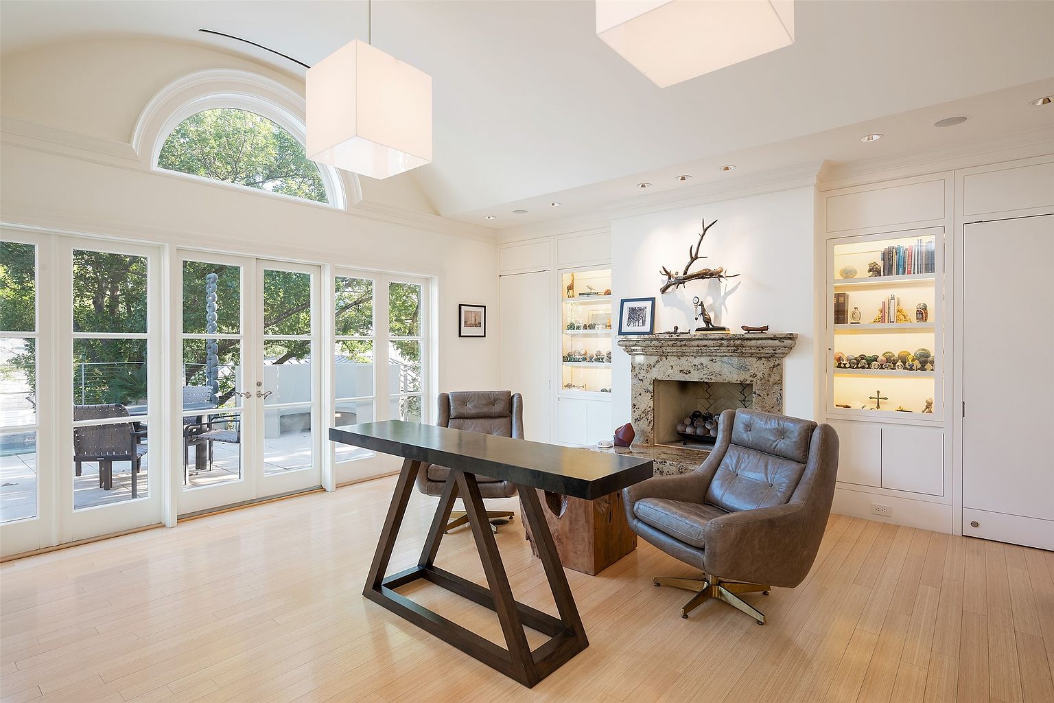 This sophisticated home office features light wood flooring and a vaulted ceiling with two large, square pendant lights. A dark, geometric-based desk sits centrally in the room, flanked by two leather swivel chairs, while a stone-clad fireplace with a decorative mantel serves as a focal point. Built-in cabinetry with illuminated shelving displays various collectibles, and a series of glass-paned doors with a large arched transom window provide abundant natural light and a view of the outdoor patio.