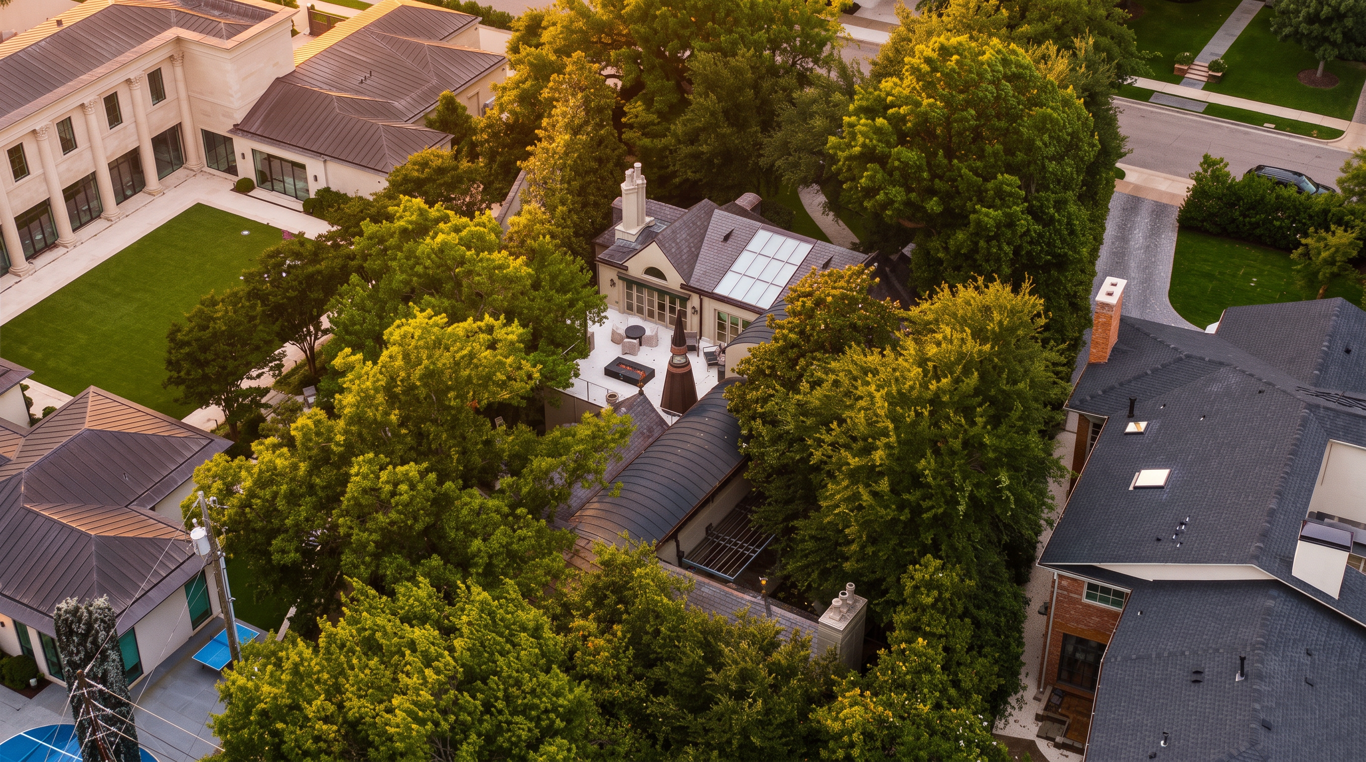 This high-angle aerial view captures a lush, tree-filled residential property featuring a complex roofline with dark metal and shingle materials. A central courtyard area includes a patio with outdoor seating and a fire feature, nestled between multiple building wings. The perspective highlights the dense landscaping and the architectural integration of the home within its private, wooded lot.