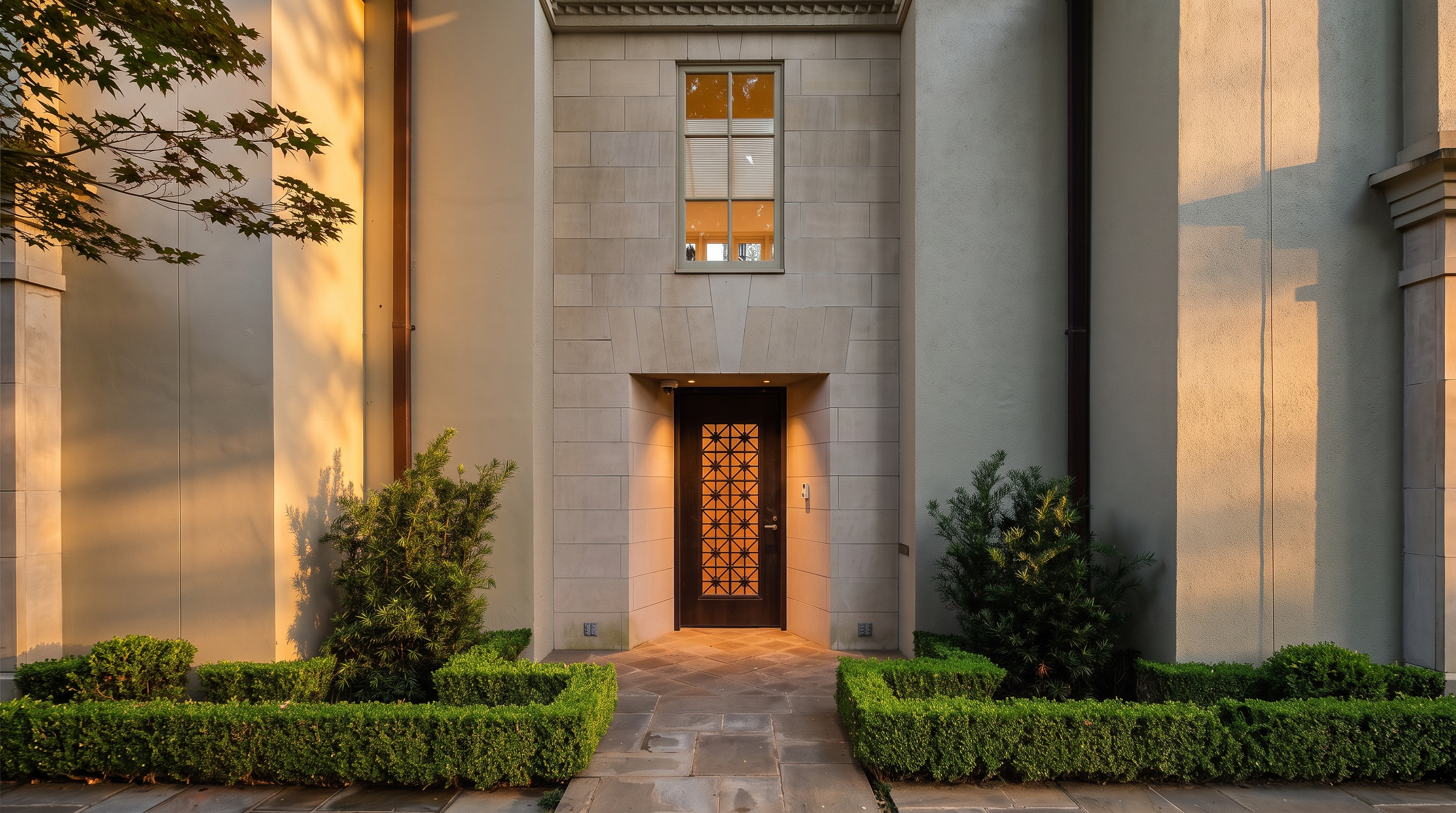 This elegant entryway features a dark, decorative metal-grille door set within a recessed alcove of light-colored stone blocks. The symmetrical facade is flanked by manicured boxwood hedges and lush greenery, creating a formal and inviting approach. A tall, multi-pane window above the door adds architectural interest, while warm lighting highlights the textured stone and entryway details.