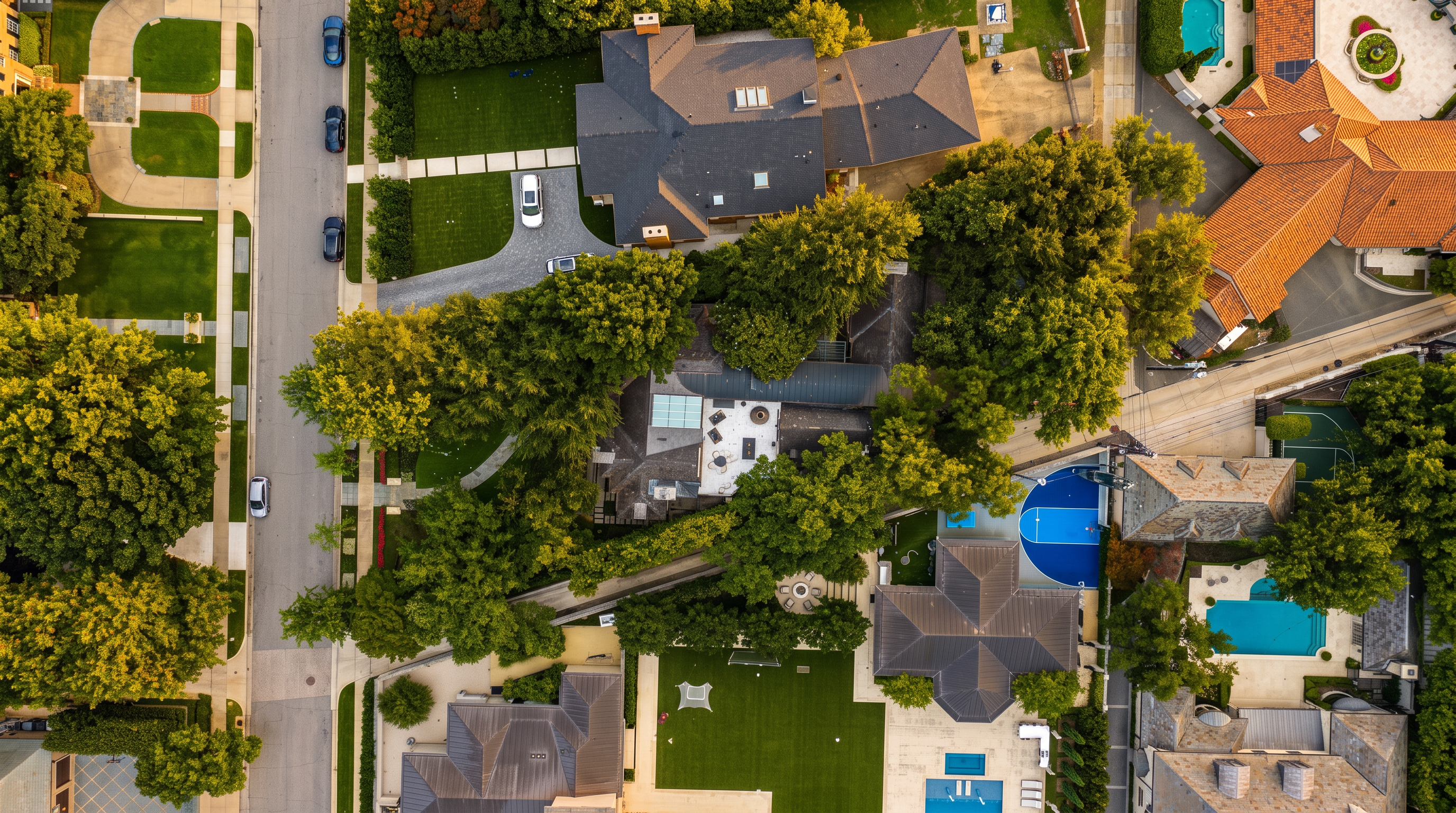This high-altitude aerial view captures a dense, upscale residential neighborhood characterized by large, multi-story homes with complex rooflines and expansive private grounds. The scene features a mix of manicured lawns, mature trees, private swimming pools, and a distinct blue-surfaced sports court, showcasing a high-density luxury living environment. The perspective provides a comprehensive layout of the properties, driveways, and surrounding street infrastructure.