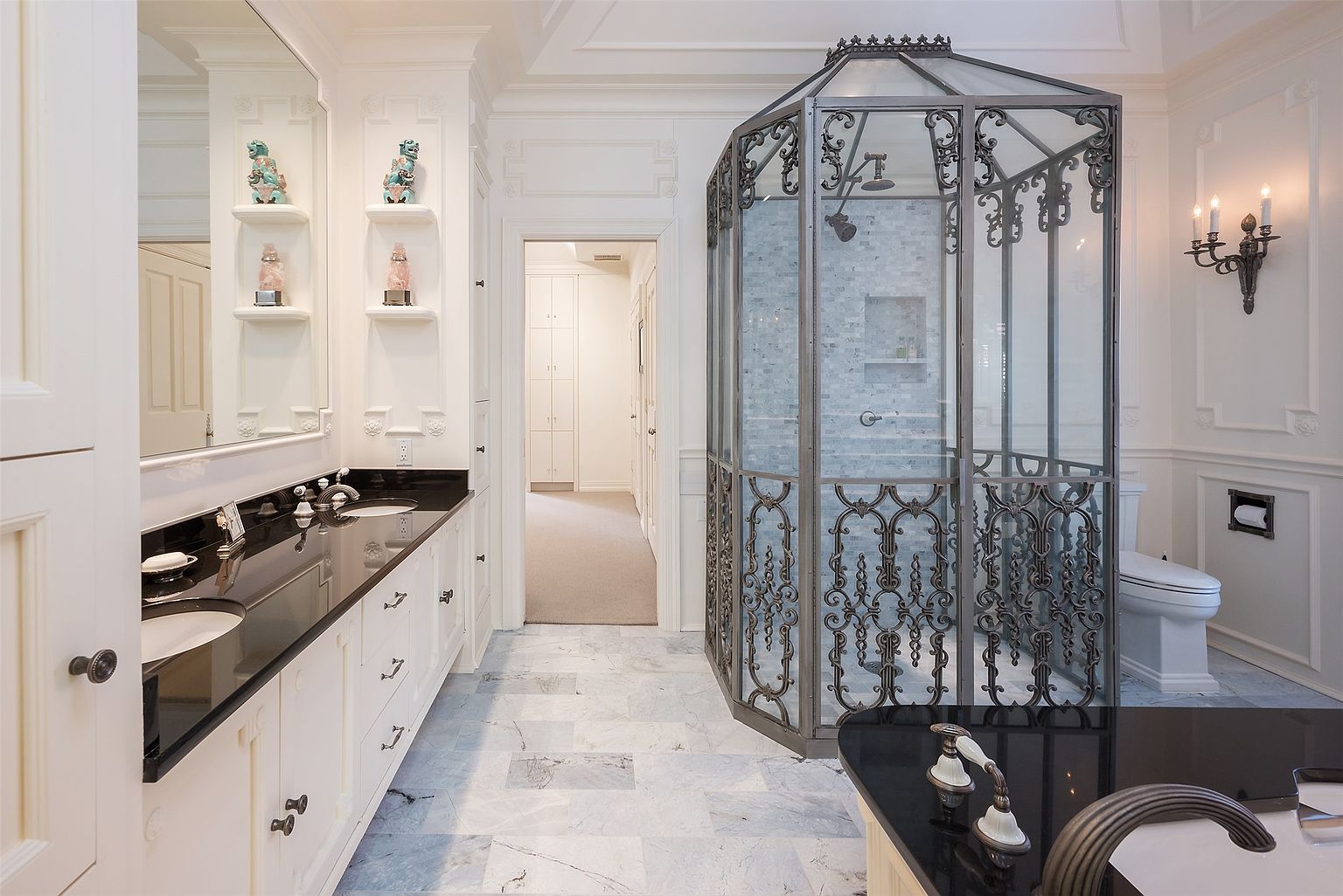 This luxurious primary bathroom features a striking custom shower enclosure with ornate metal detailing and glass panels. The space is anchored by a double vanity with a dark stone countertop and white cabinetry, complemented by light-toned stone flooring. A glimpse into an adjacent hallway reveals a well-organized storage area, while wall-mounted sconces add a touch of classic elegance to the bright, sophisticated room.
