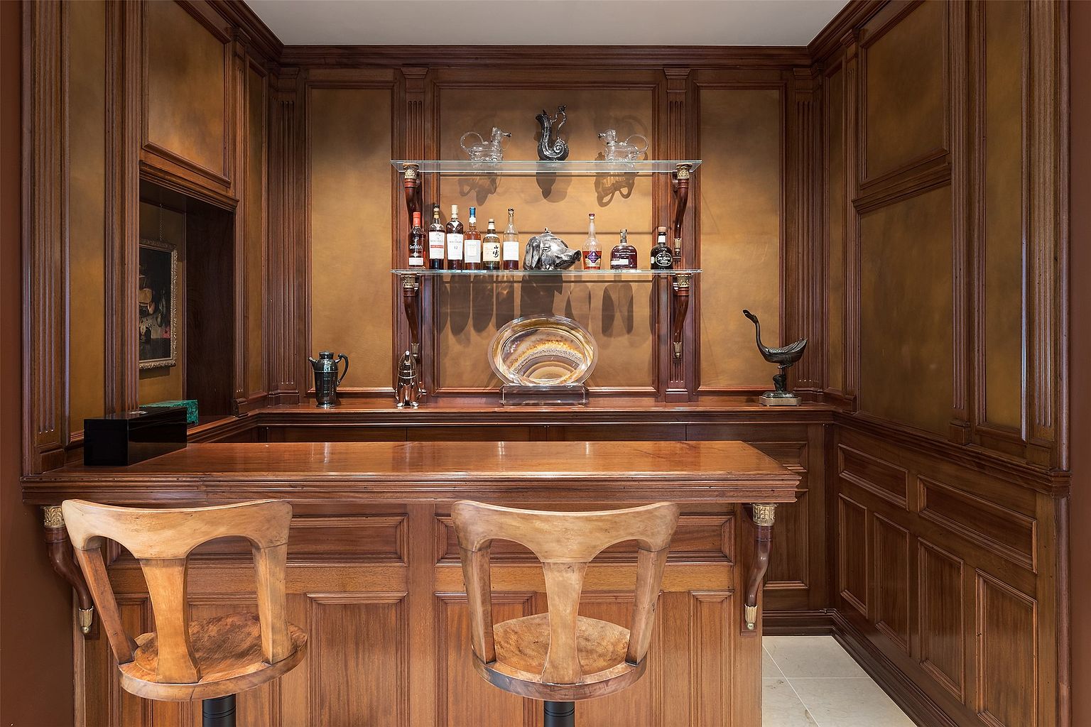 This image showcases a sophisticated home bar area characterized by rich, dark wood paneling and cabinetry that creates a classic, club-like atmosphere. The bar features a polished wood countertop, two wooden stools, and two glass shelves displaying a collection of spirits and decorative objects. The warm, ambient lighting highlights the textured wall finish and the intricate woodwork, providing an elegant space for entertaining.