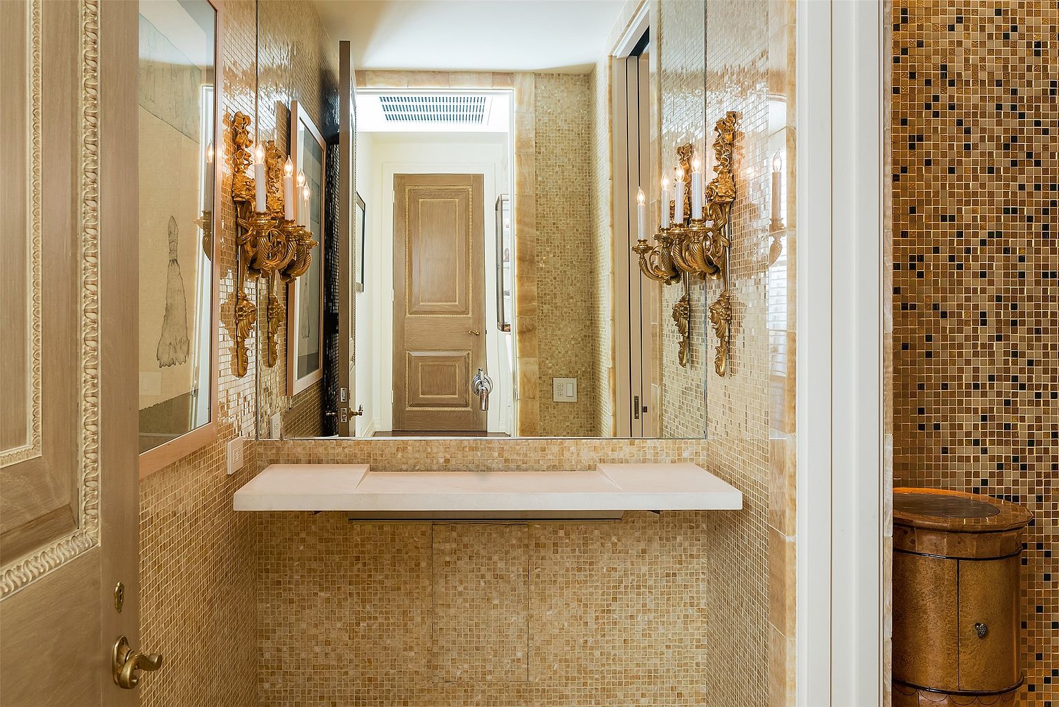 This elegant powder room features intricate gold-toned mosaic tile walls and a minimalist stone vanity shelf. Ornate gold wall sconces flank a large mirror, reflecting the hallway and a closed wooden door beyond. The space exudes a luxurious, classic aesthetic with a focus on rich textures and warm lighting.