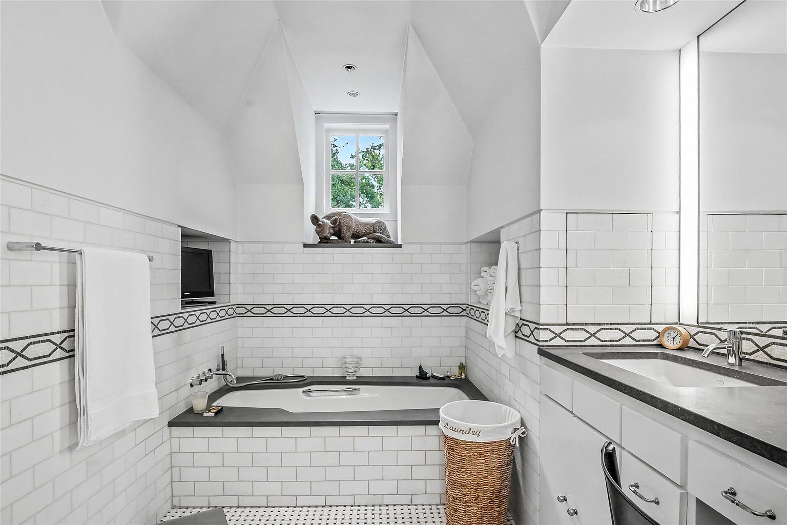 This bright and classic bathroom features a deep soaking tub set into a dark stone surround, accented by white subway tile walls with a decorative patterned border. A window centered above the tub brings in natural light, while a vanity with a matching dark stone countertop sits to the right. The space is finished with a woven laundry basket and clean, white cabinetry, creating a serene and timeless atmosphere.