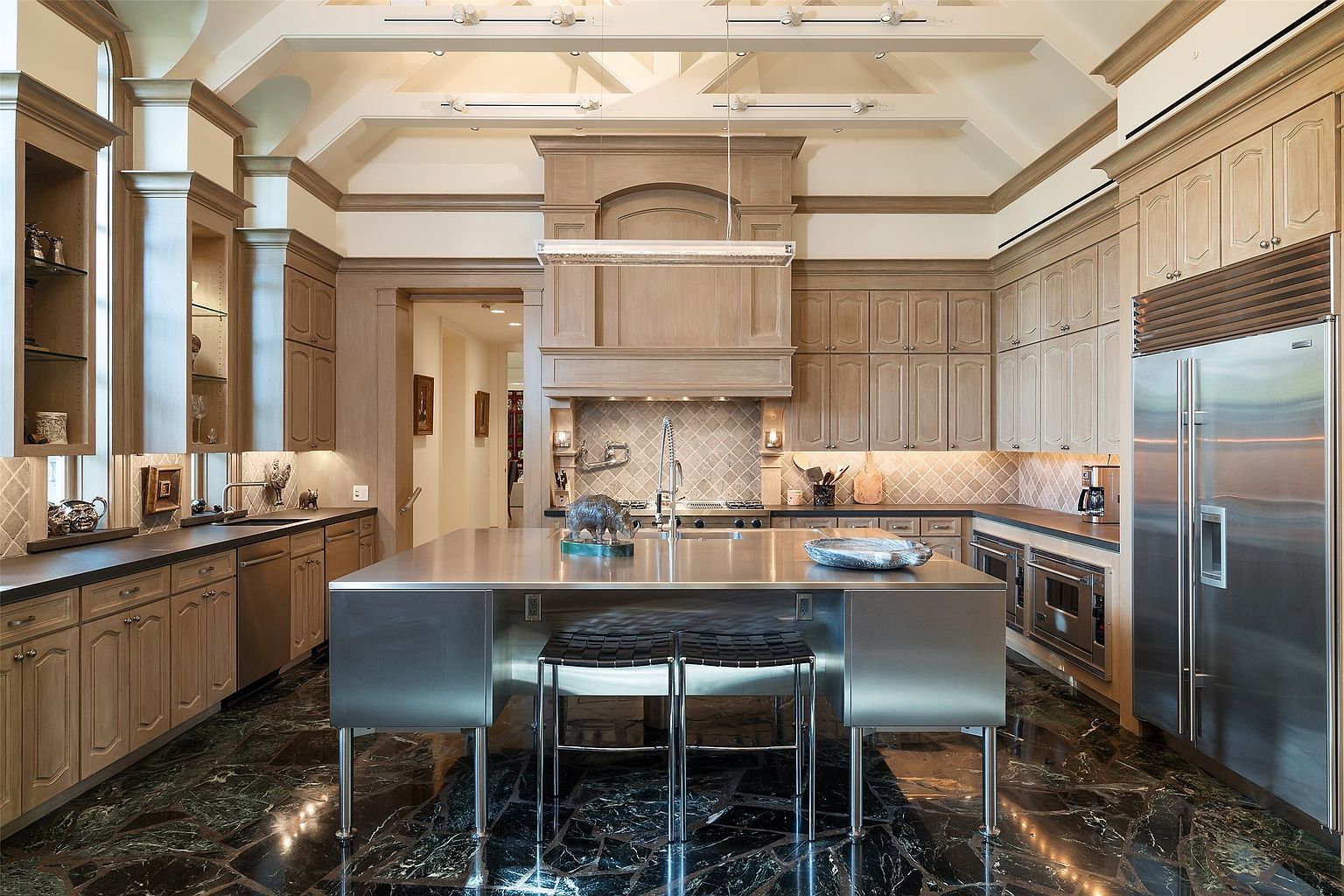 This spacious, high-ceilinged kitchen features a blend of traditional light wood cabinetry and a modern, industrial-style stainless steel central island. The room is anchored by a striking dark, veined stone floor and a custom range hood that serves as a focal point above the stove. The perspective is a centered, eye-level shot that highlights the symmetry of the cabinetry and the contrast between the warm wood tones and the cool metallic surfaces.