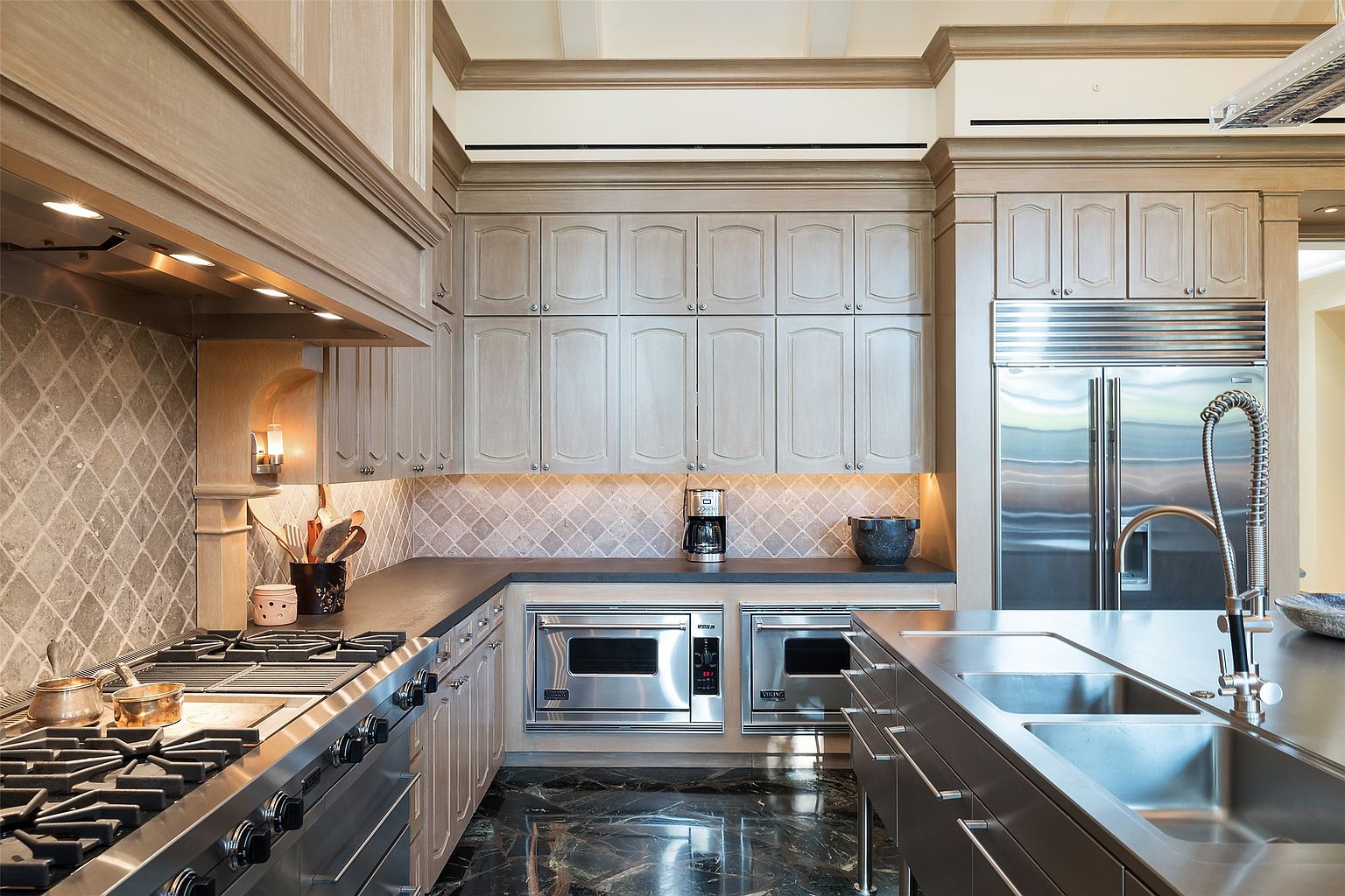 This high-end kitchen features light wood cabinetry, dark stone countertops, and a professional-grade stainless steel range and refrigerator. A diamond-patterned tile backsplash adds texture, while the dark, polished stone flooring provides a sophisticated contrast. The perspective captures the functional layout, highlighting the integrated appliances and the central island with a stainless steel sink.