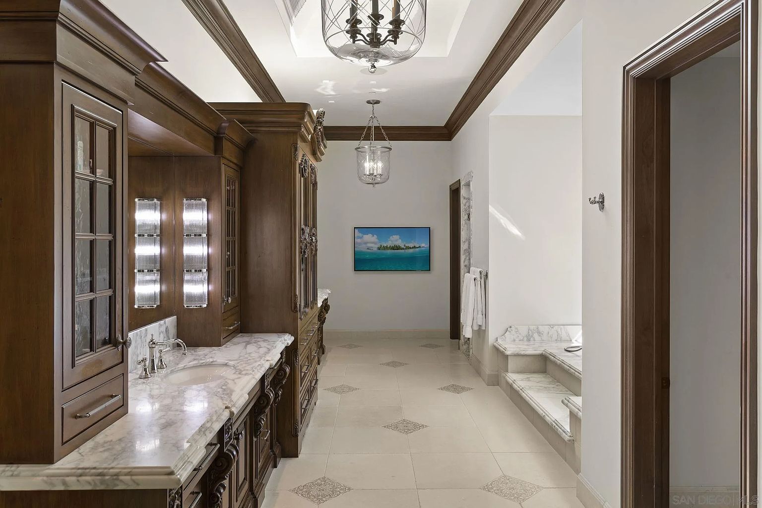 This is a luxurious primary bathroom featuring custom dark wood cabinetry with marble countertops and integrated lighting. The long vanity provides ample storage and counter space, while the tile flooring adds an elegant touch. A large television is mounted on the wall at the end of the hallway, and the room is illuminated by decorative pendant lights and recessed lighting.