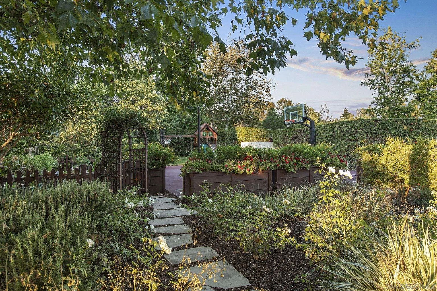 This image showcases a beautifully landscaped backyard garden. A stone pathway leads through lush greenery and flower beds, with a wooden arbor and fence adding rustic charm. In the background, a basketball hoop and play structure suggest family-friendly amenities, all framed by mature trees and hedges, creating a private and inviting outdoor space.
