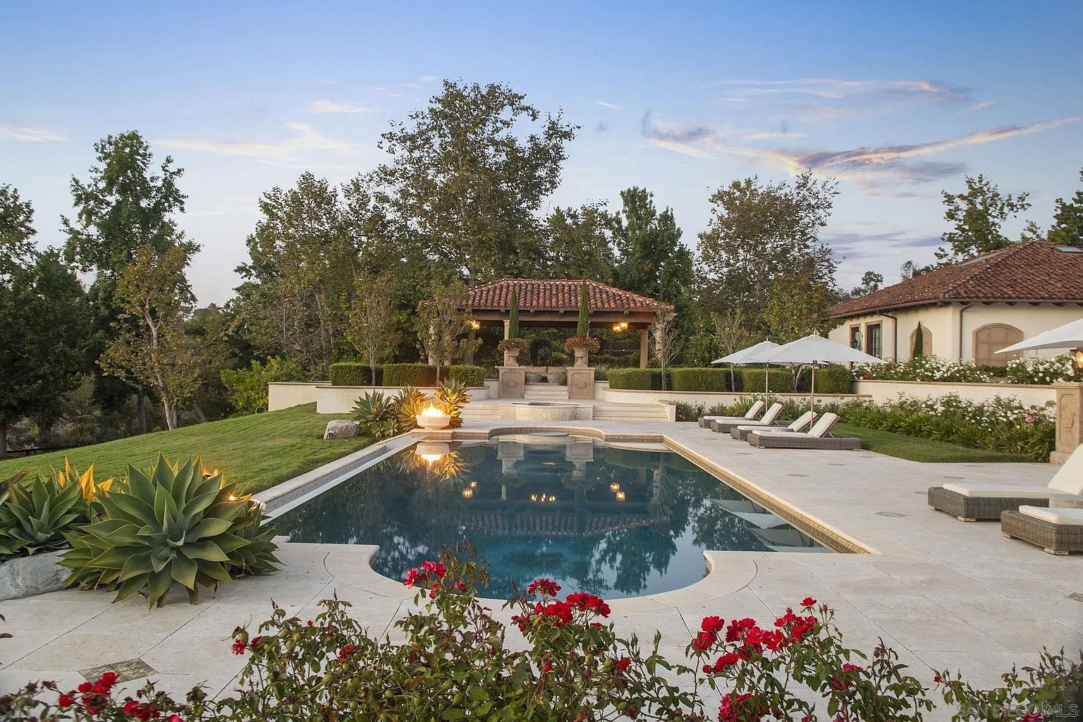This image showcases a luxurious backyard pool and spa area. The pool features a unique shape and is surrounded by a stone patio, lounge chairs, and lush landscaping, including red roses and agave plants. A covered gazebo sits at the far end of the pool, adding an element of elegance and relaxation to the outdoor space. The house is visible in the background, suggesting a private and upscale setting.