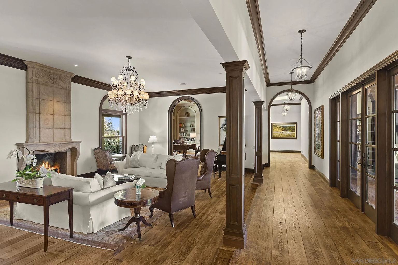 This is an interior shot of a luxurious living room featuring hardwood floors, a stone fireplace, and elegant furnishings. The room is bathed in natural light, highlighting the detailed architectural features such as the dark wood trim and arched doorways. A grand chandelier adds a touch of opulence to the space.