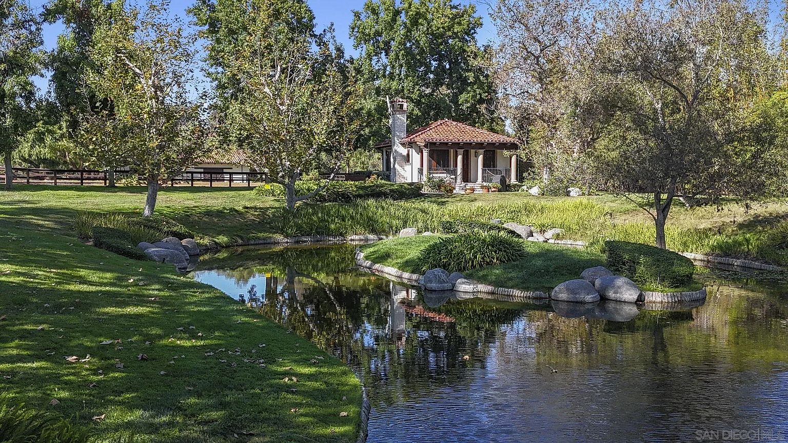 This exterior shot showcases a serene yard and garden featuring a tranquil pond with lush greenery and decorative rocks. A charming building with a red tile roof and columned porch sits in the background, adding to the property's appeal. The scene evokes a sense of peacefulness and natural beauty, highlighting the outdoor living space.