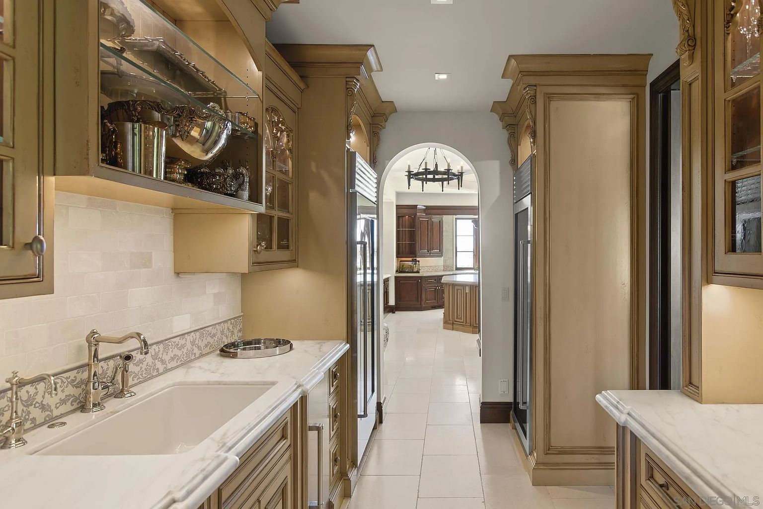 This is an interior shot of a luxurious kitchen hallway. The hallway features custom cabinetry with glass displays, marble countertops, and high-end appliances. The arched doorway leads to another part of the kitchen, showcasing the home's open layout and sophisticated design.