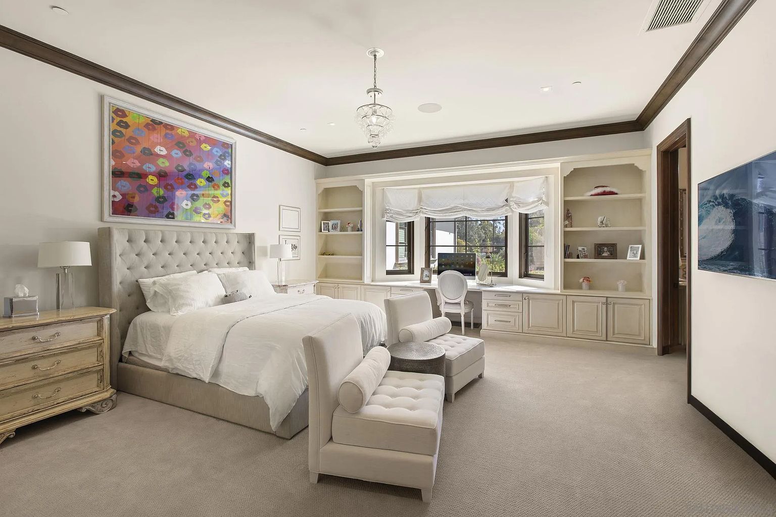 This is a luxurious primary bedroom featuring a large bed with a tufted headboard, white linens, and two lounge chairs. The room has built-in shelving and a desk area by the window, providing ample storage and workspace. An eye-catching piece of art hangs above the bed, adding a pop of color to the otherwise neutral palette.