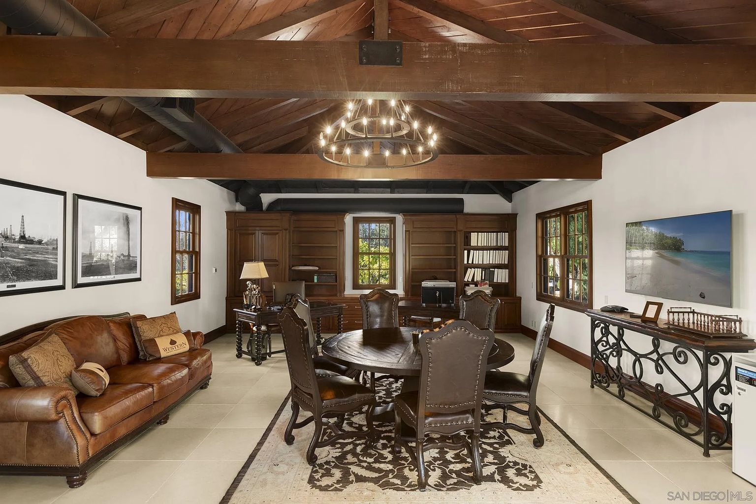 This is an interior shot of a luxurious office or study. The room features a large wooden built-in bookcase, a round wooden table with leather chairs, and a leather sofa. The ceiling is wooden with exposed beams and a chandelier, creating a warm and inviting atmosphere.