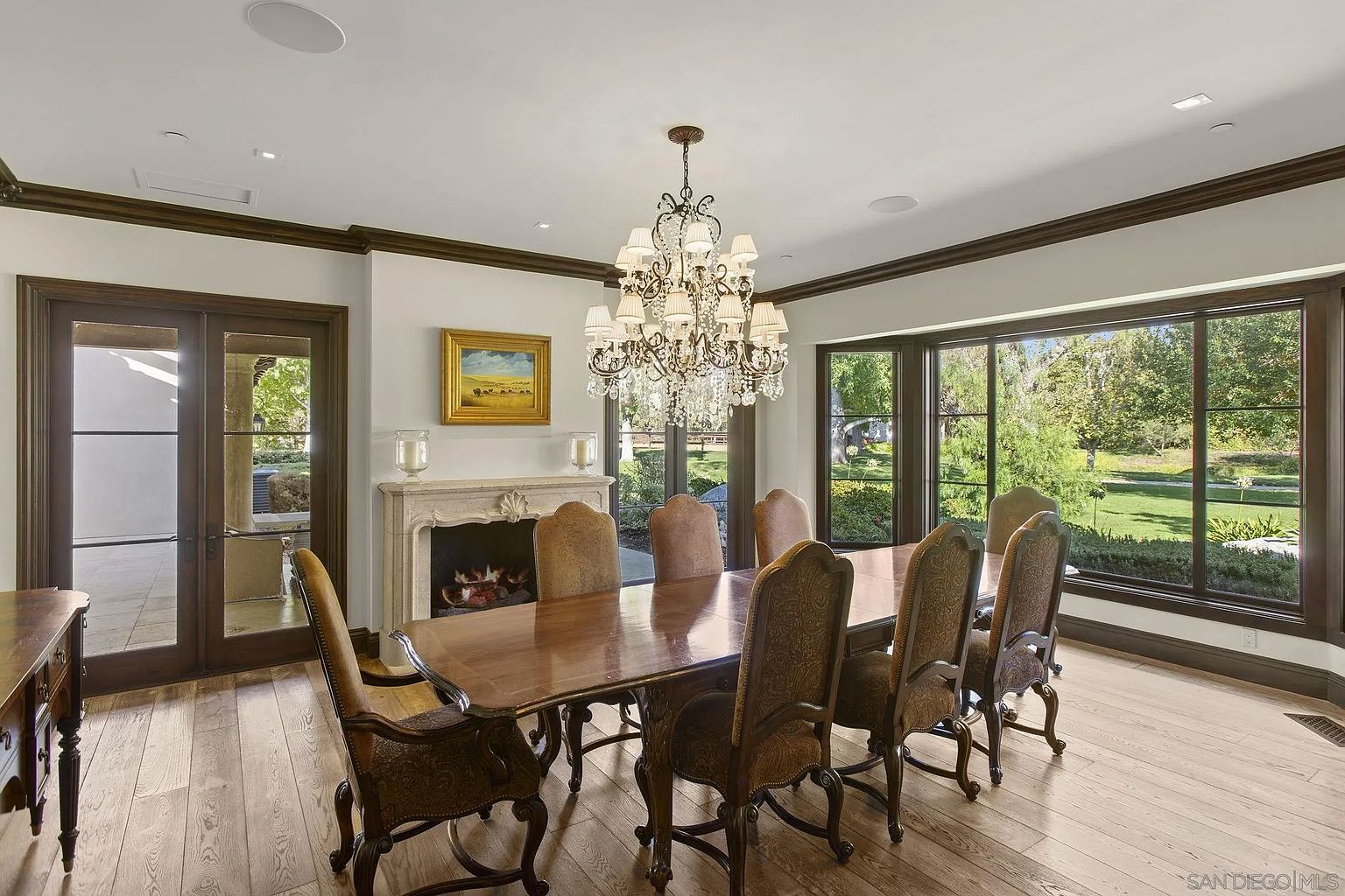 This is an interior shot of a formal dining room. The room features a large wooden dining table with upholstered chairs, a decorative chandelier, and a fireplace. Large windows provide natural light and a view of the exterior, enhancing the room's elegant and inviting atmosphere.
