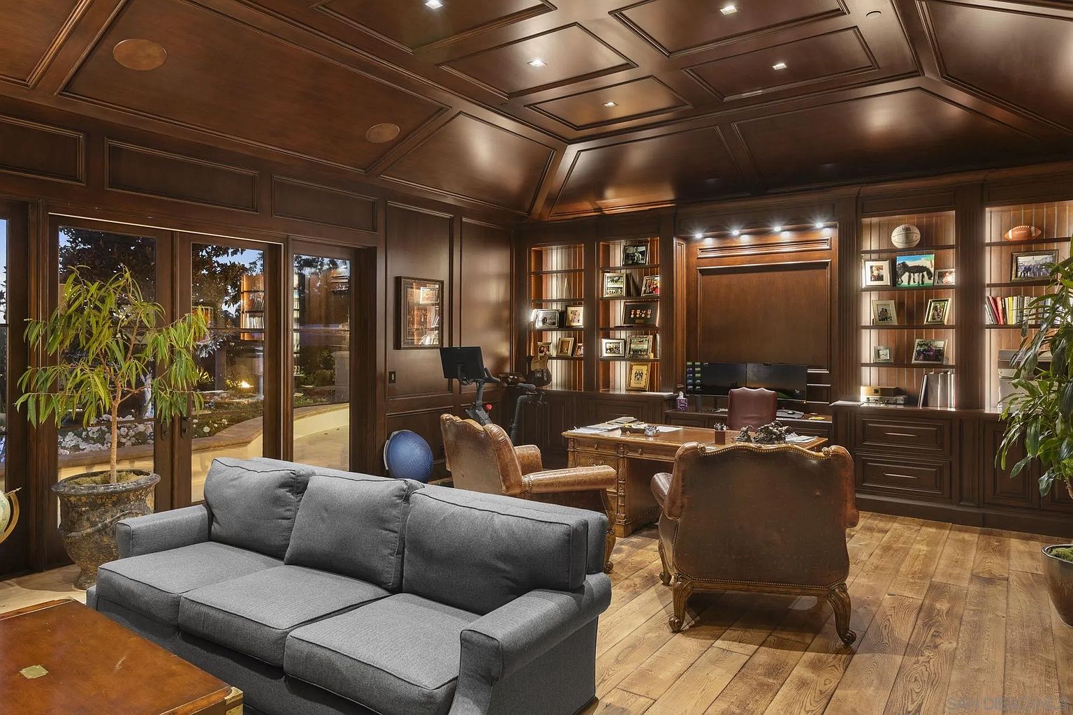 This is a luxurious home office featuring rich wood paneling and built-in bookshelves filled with decorative items and personal photos. A large wooden desk is the focal point, complemented by leather chairs and a comfortable gray sofa. The room exudes a sense of sophistication and warmth, perfect for a professional or personal workspace.