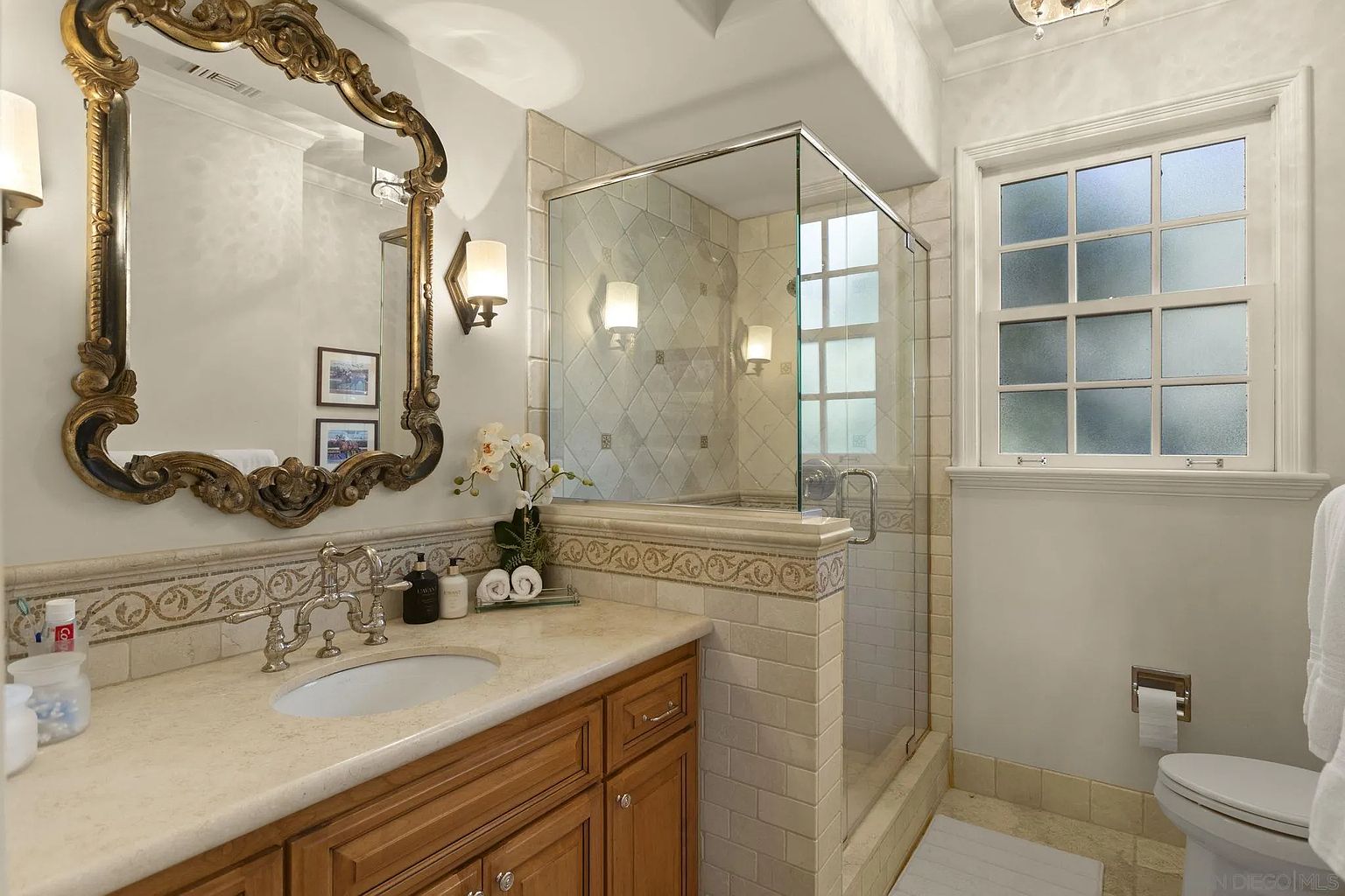 This bathroom features a vanity with a light-colored countertop and wooden cabinets, complemented by an ornate gold-framed mirror. A glass-enclosed shower with tiled walls is adjacent to the vanity, and a window provides natural light. The overall aesthetic is traditional and well-maintained.