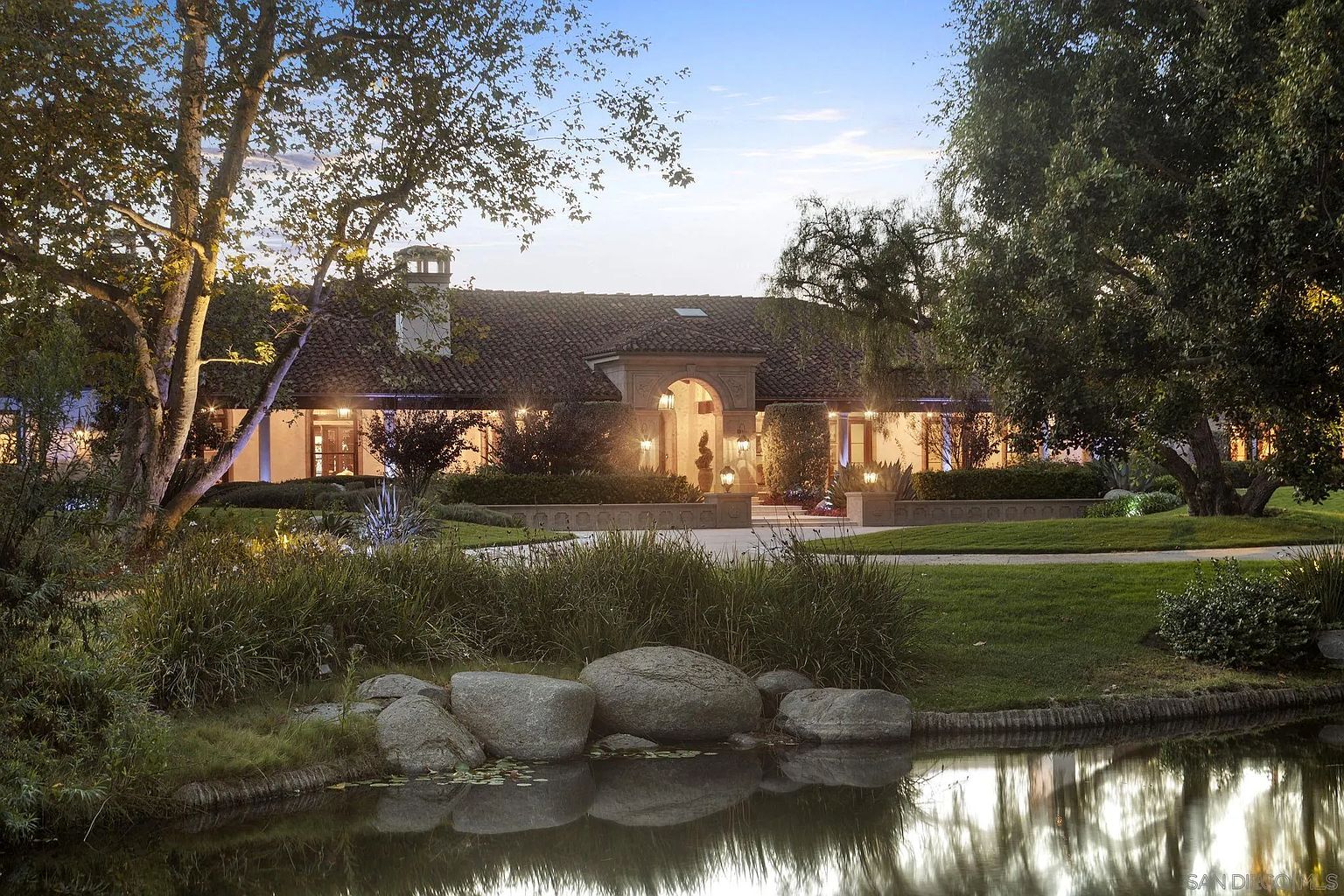 This is a front view of a luxurious estate, showcasing a grand entrance with an arched doorway and meticulously landscaped grounds. The property features a tiled roof, elegant lighting, and a serene pond in the foreground, creating a picturesque and inviting scene. Mature trees frame the house, adding to the sense of privacy and established beauty.