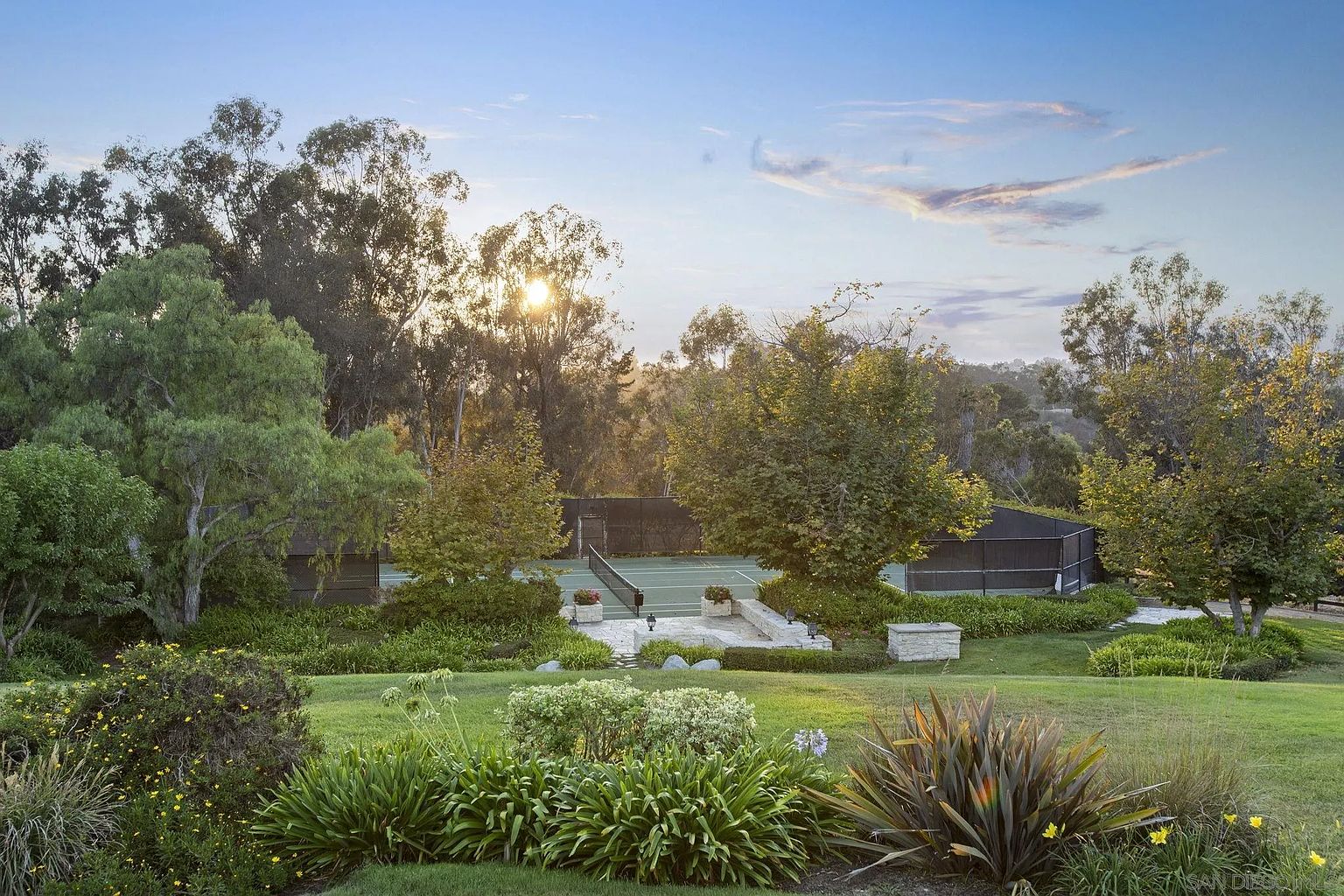 This exterior shot showcases a beautifully landscaped yard and garden, featuring lush green lawns, mature trees, and meticulously arranged flower beds. A tennis court is visible in the background, suggesting a property with ample space and recreational amenities. The overall impression is one of tranquility and well-maintained elegance.