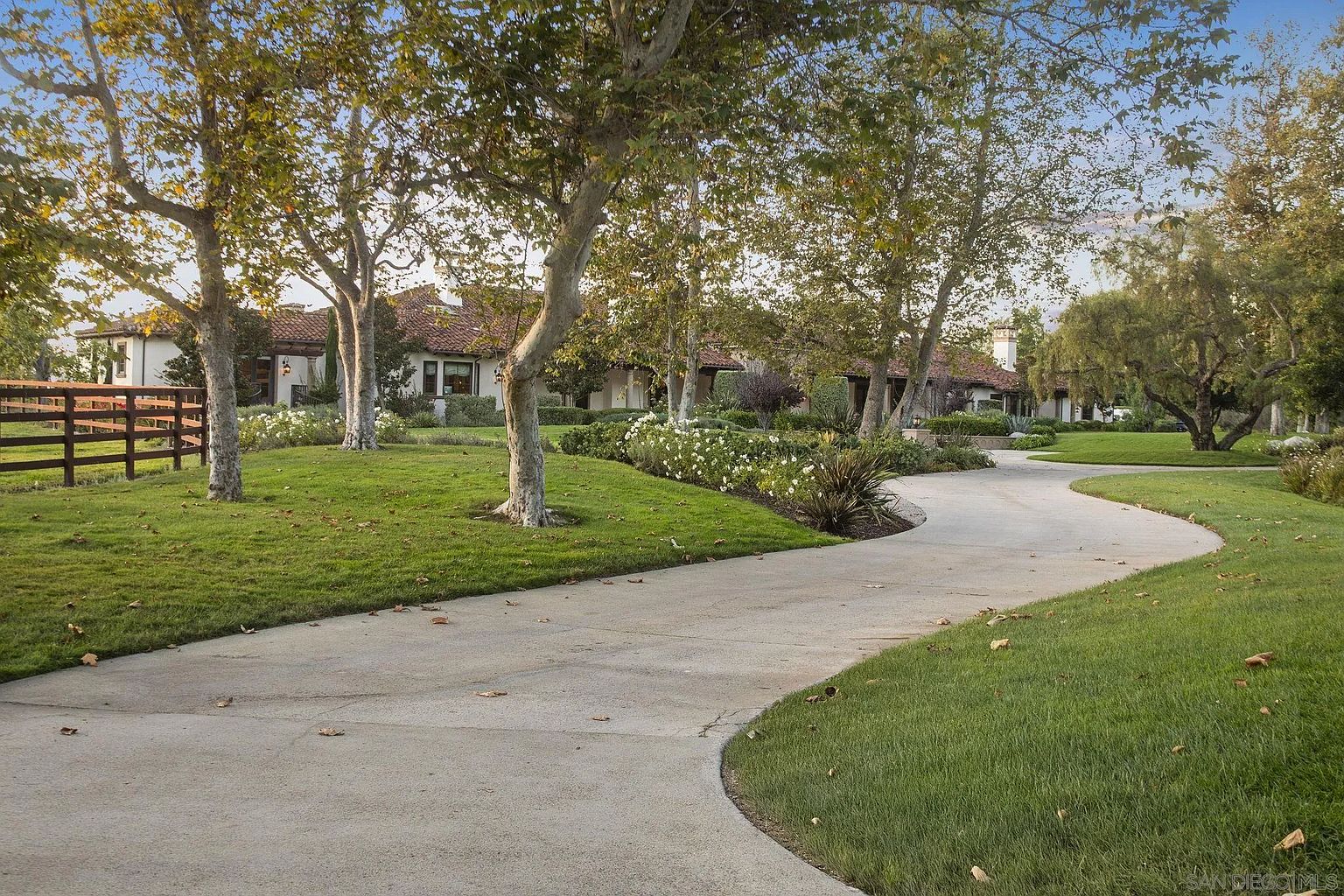 This image showcases a beautifully landscaped yard with a winding driveway leading to a large home. Mature trees line the driveway and frame the house, while manicured lawns and flower beds add to the property's curb appeal. A wooden fence is visible on the left, suggesting a well-maintained and private estate.