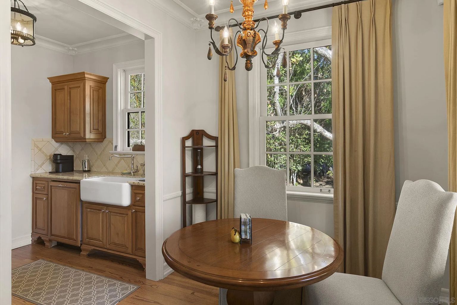 This inviting dining area features a round wooden table set beneath an ornate chandelier, complemented by two upholstered chairs. A window with a view of lush greenery is framed by elegant drapes, adding natural light and warmth to the space. The adjacent kitchen area is visible through an archway, showcasing wooden cabinetry and a farmhouse sink.