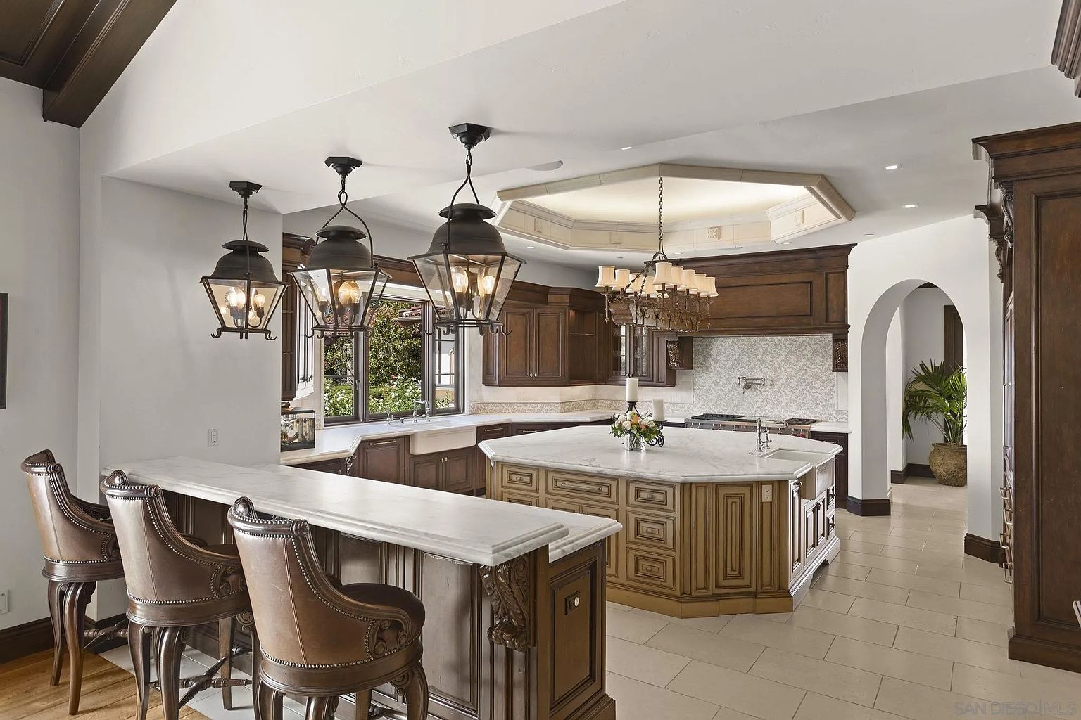 This is a spacious and luxurious kitchen featuring dark wood cabinetry, marble countertops, and a large central island. The kitchen is well-lit with a combination of pendant lights and a chandelier, creating a warm and inviting atmosphere. The design incorporates traditional elements with modern functionality, making it ideal for both cooking and entertaining.