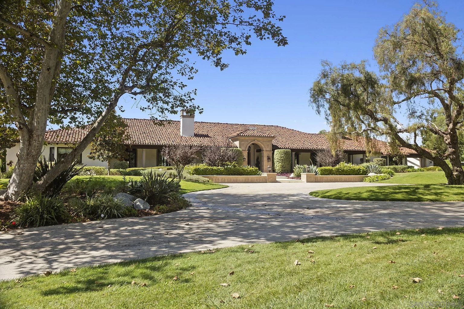 This is a front view of a luxurious single-story home with a red tile roof and a grand arched entryway. The property features a circular driveway, lush green lawns, and mature trees that frame the house, creating a sense of privacy and elegance. The landscaping is meticulously maintained, adding to the overall curb appeal.