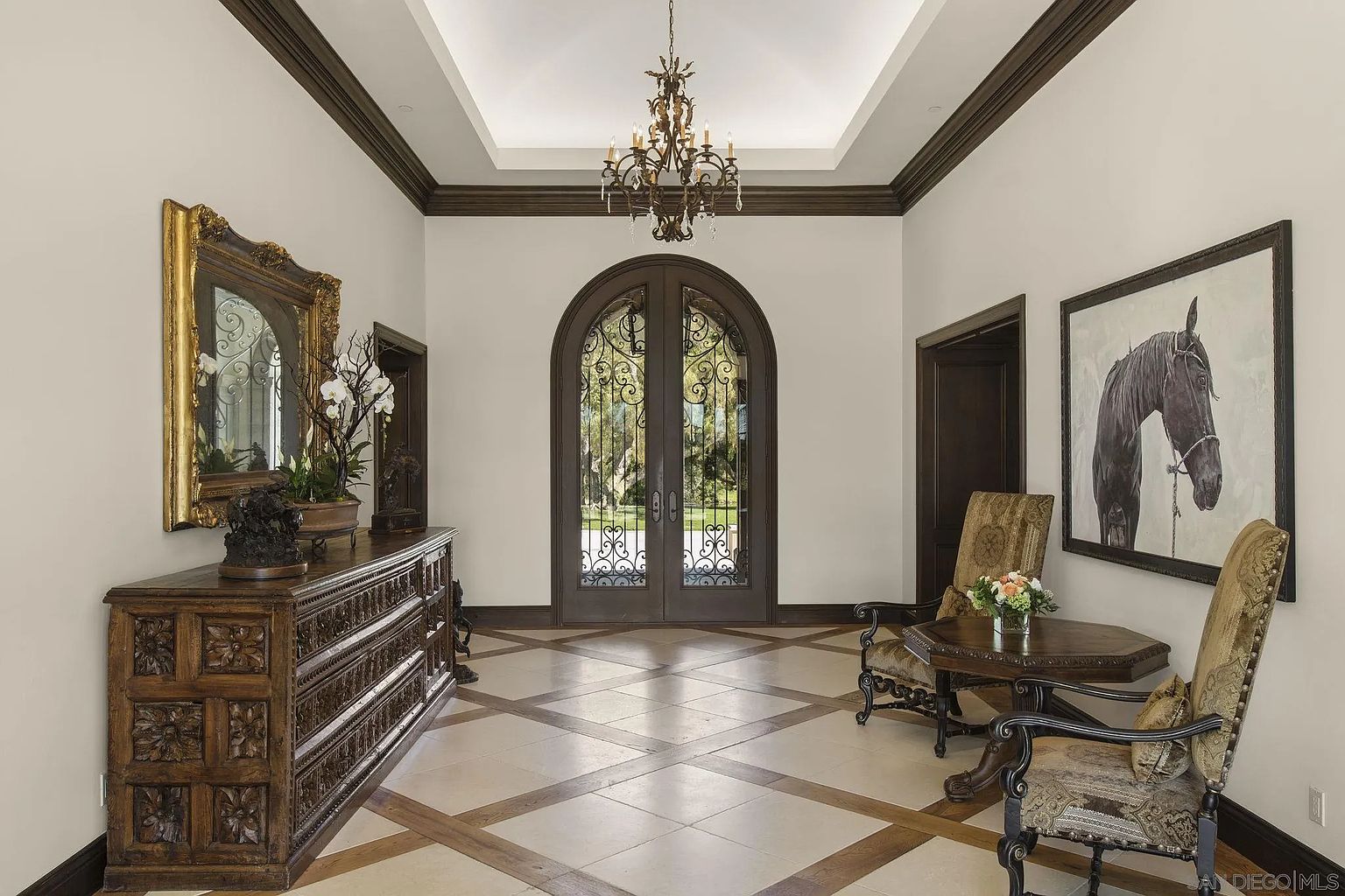 This grand foyer features a striking combination of classic and elegant design elements. The arched double doors with intricate ironwork provide a view to the exterior, while a large ornate mirror and a horse painting adorn the walls. The patterned tile floor and the chandelier add to the luxurious feel of the space.