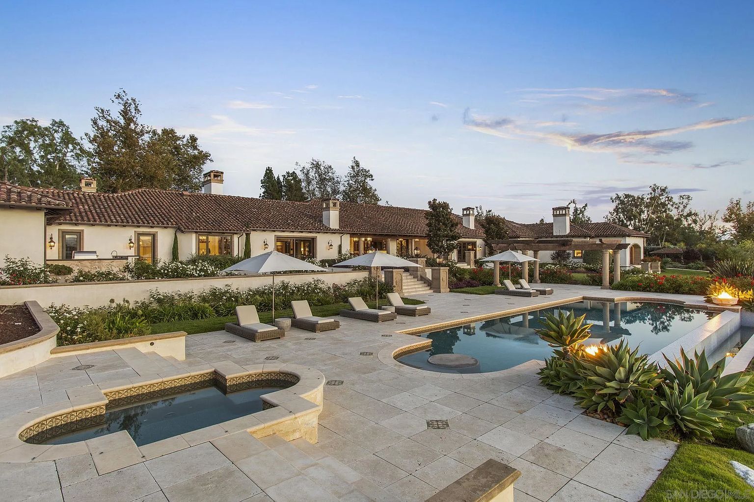 This image showcases a luxurious backyard featuring a large swimming pool and spa area. The pool has a unique shape with a built-in seating area, surrounded by lush landscaping and elegant patio furniture. The property's architecture is visible in the background, suggesting a high-end estate with a focus on outdoor living and entertainment.