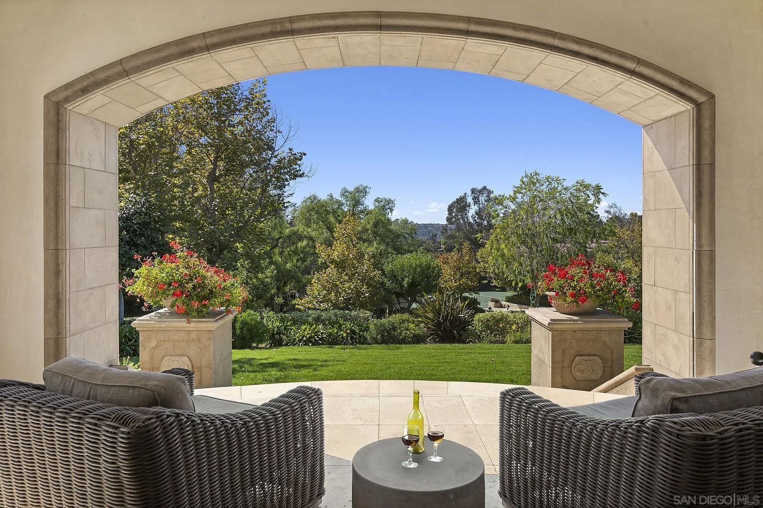 This image showcases a luxurious outdoor patio area, framed by an elegant stone archway. Two wicker chairs flank a small round table with a bottle of wine and two glasses, suggesting a relaxing and inviting space. Beyond the patio, a meticulously maintained lawn and lush garden create a serene backdrop, enhancing the property's appeal.