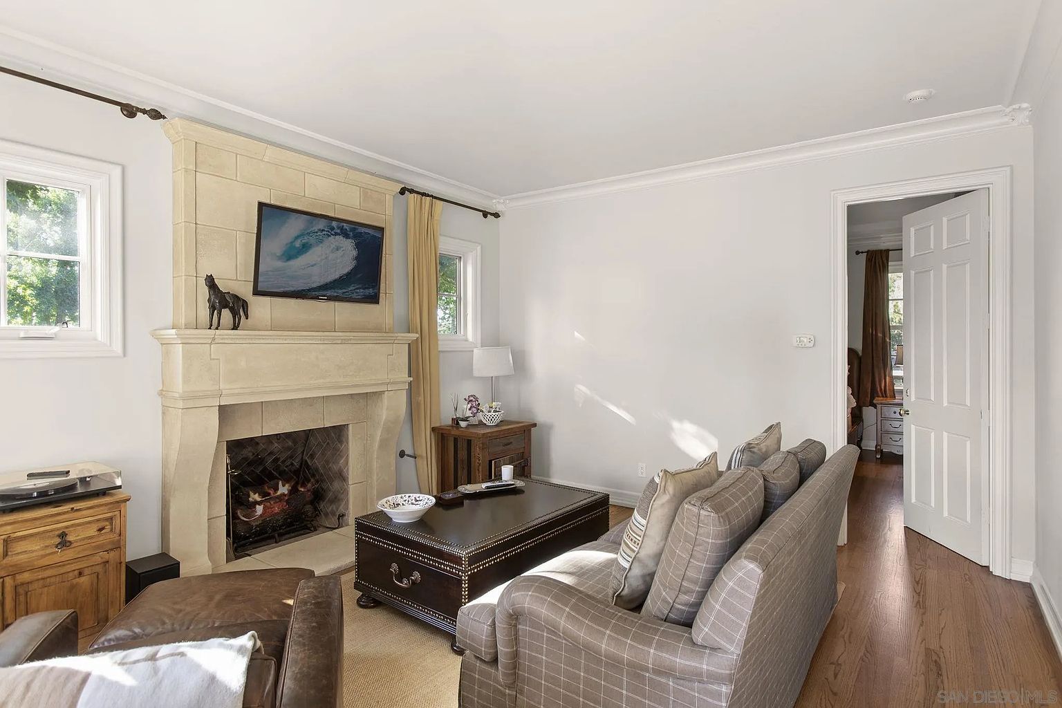 This is an interior shot of a living room featuring a stone fireplace with a mounted television above it. A plaid sofa faces the fireplace, with a dark wood coffee table in between. The room has hardwood floors and neutral-colored walls, creating a cozy and inviting atmosphere.