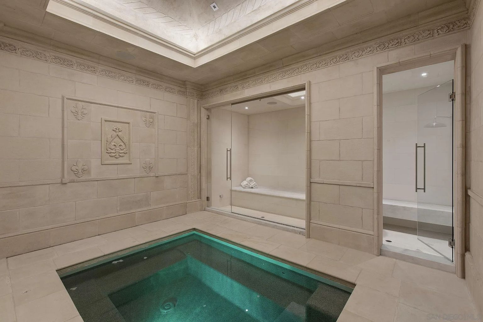 This is a luxurious primary bathroom featuring a built-in spa or hot tub with turquoise water, a glass-enclosed steam room with a bench, and a separate glass-enclosed shower. The walls are constructed of light-colored stone blocks with decorative moldings, and the ceiling has a recessed, illuminated design, creating a serene and upscale atmosphere.