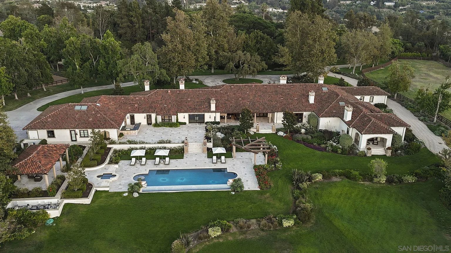 This aerial view showcases a sprawling estate with a red-tiled roof, white stucco walls, and meticulously landscaped grounds. A large swimming pool is a focal point, surrounded by patio areas and lush greenery. The property exudes luxury and privacy, nestled among mature trees and rolling hills.